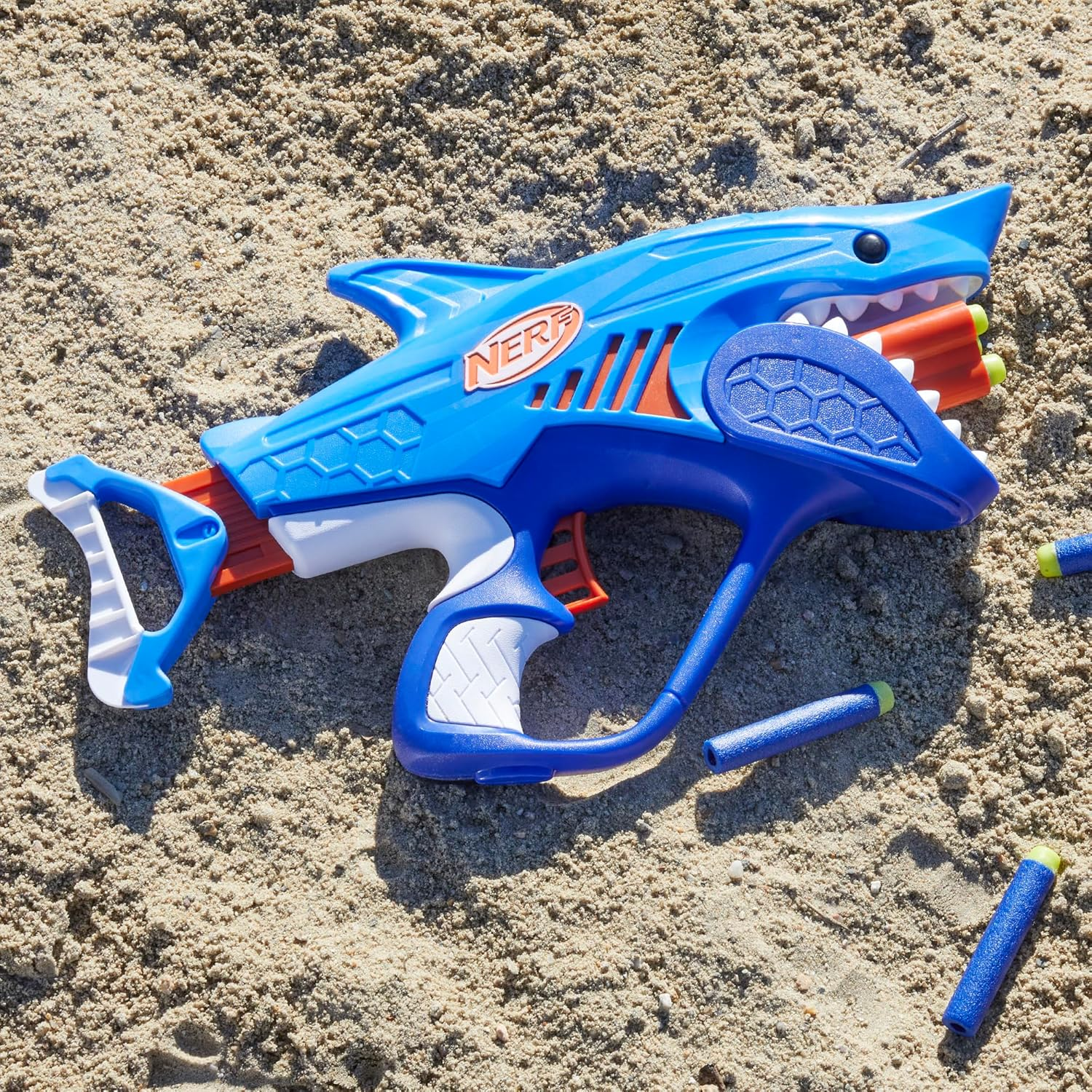 Una pistola de dardos Nerf azul con forma de tiburón yace en arena con dardos.
