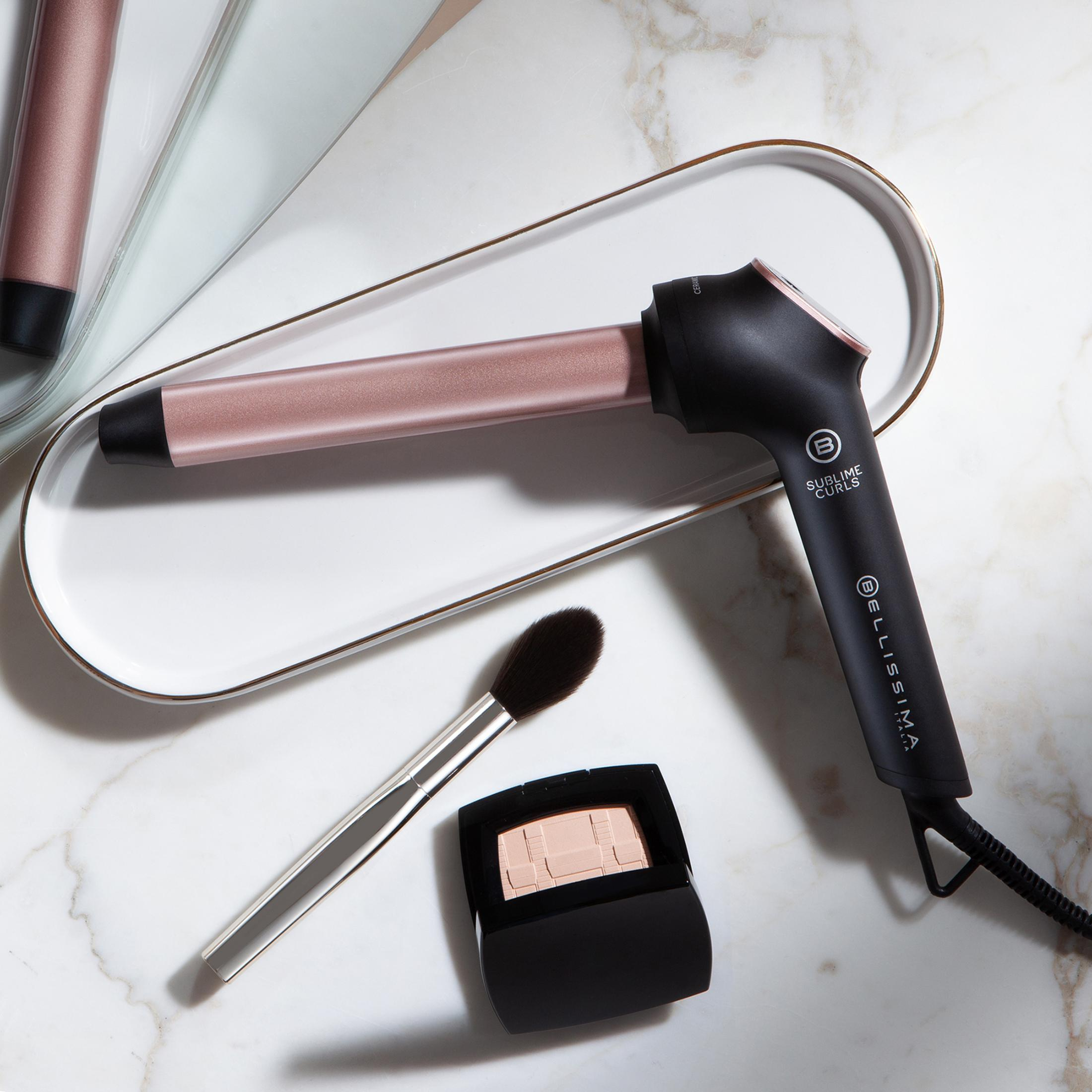 Ein Lockenstab mit Roségold-Akzenten, ein Pinsel und ein Make-up-Puder liegen auf einem Marmortisch.