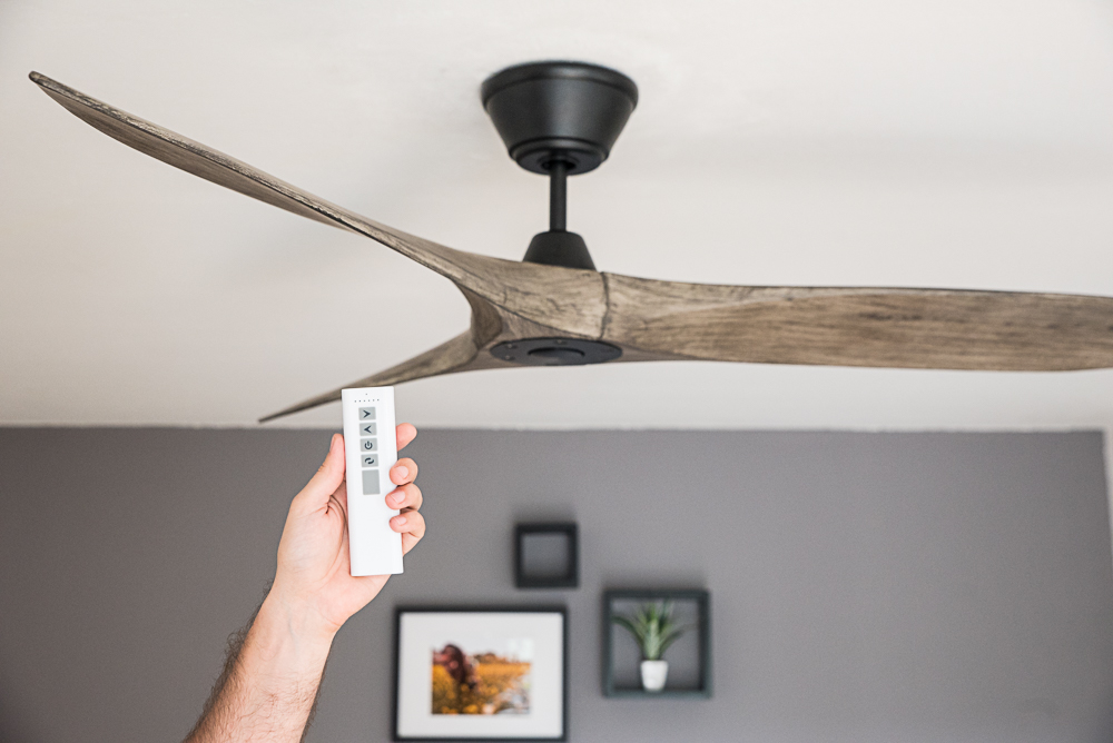 Una mano tiene un telecomando bianco, puntato verso un moderno ventilatore da soffitto in legno.