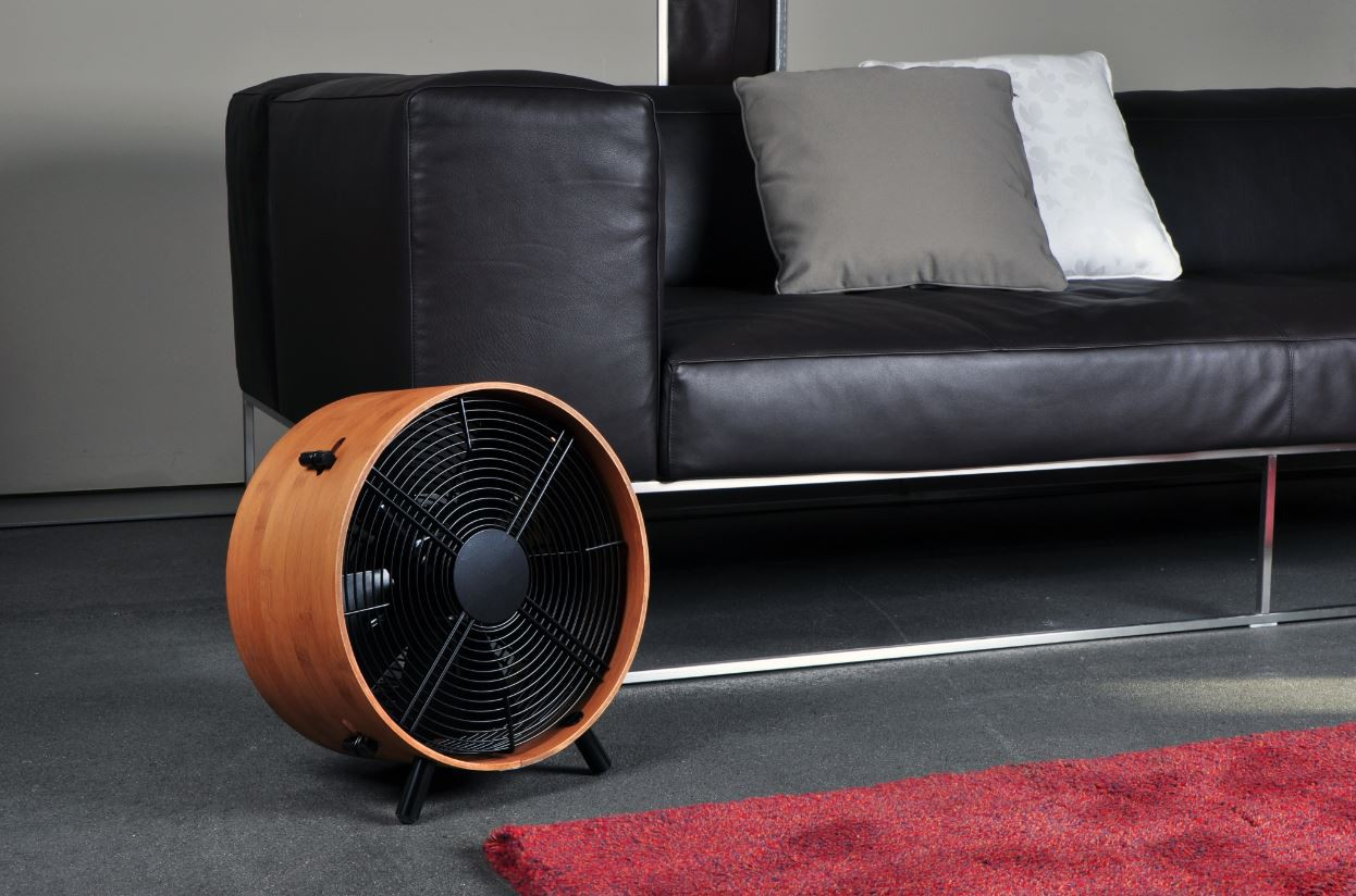 Een ronde houten ventilator staat voor een zwarte leren bank met kussens.