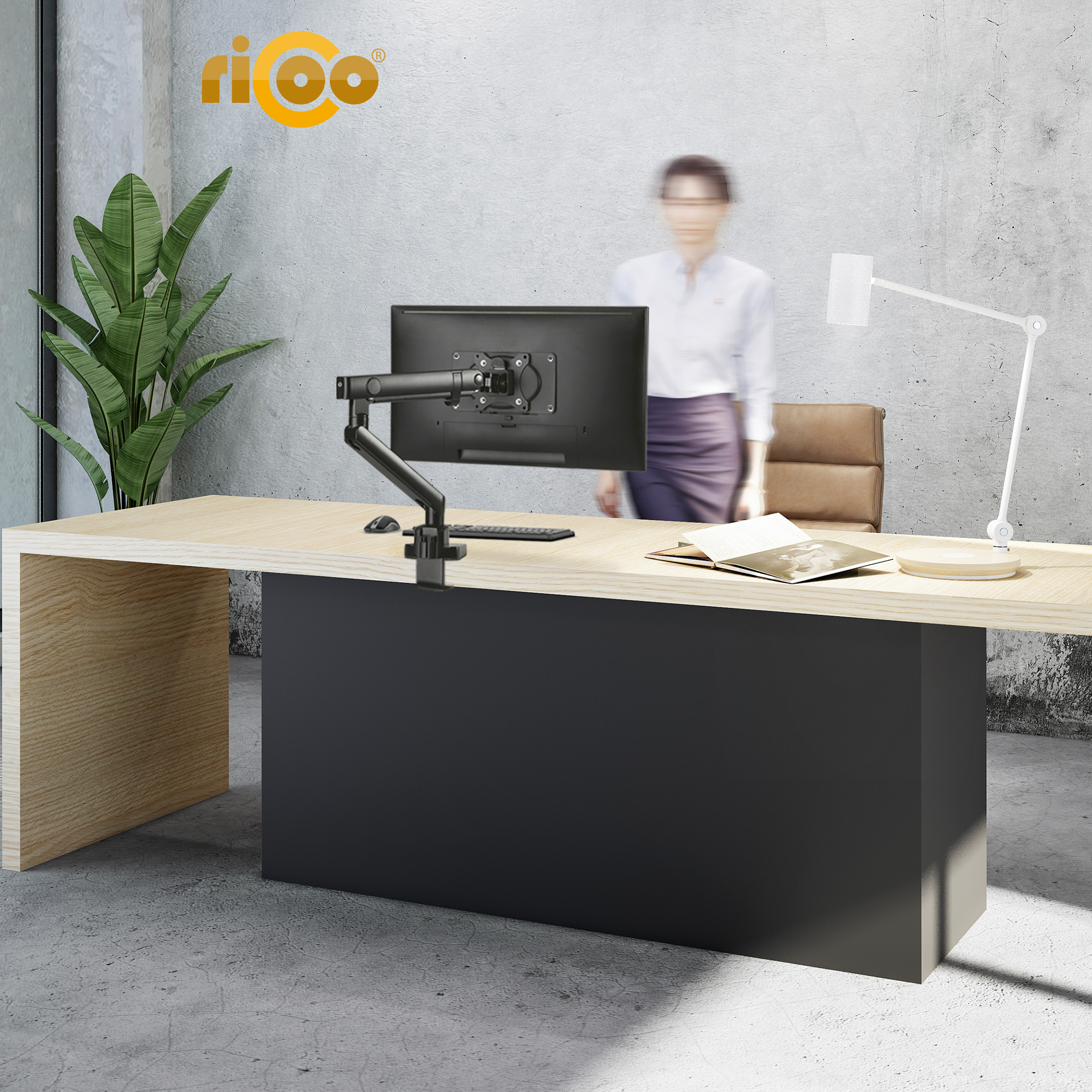 Büro mit Schreibtisch, Monitorarm, Lampe, Pflanze und Person im Hintergrund.