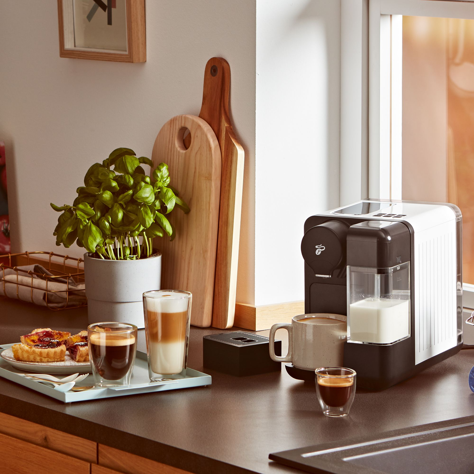 Kaffeemaschine mit Milchbehälter, Tassen, Gebäck und Pflanzen auf einer Arbeitsplatte.