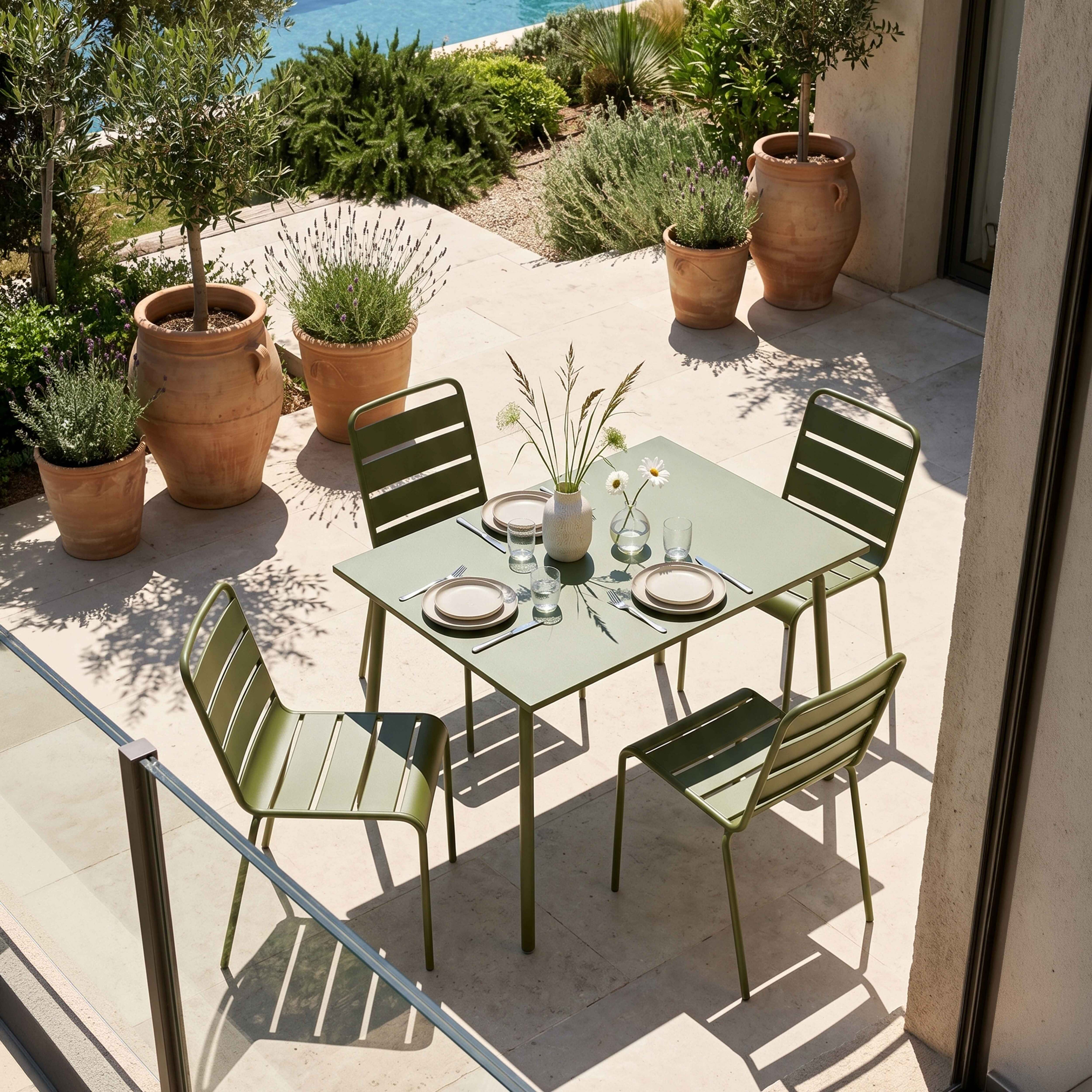Ein grüner Gartentisch mit vier Stühlen, gedeckt für eine Mahlzeit im Freien, mit Pflanzen und einem Pool im Hintergrund.