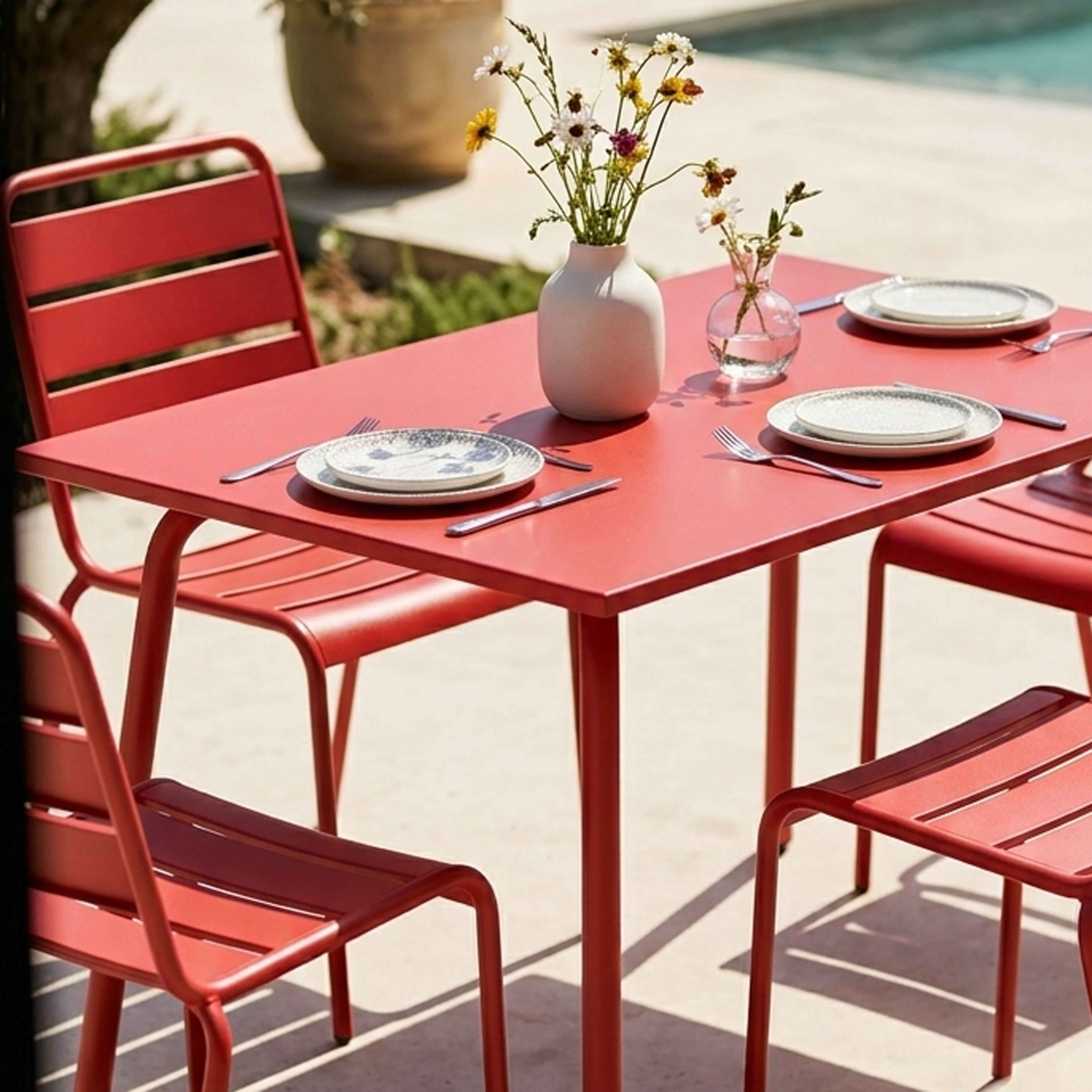 Ein roter Tisch mit Blumen und Tellern, daneben rote Stühle im Freien.