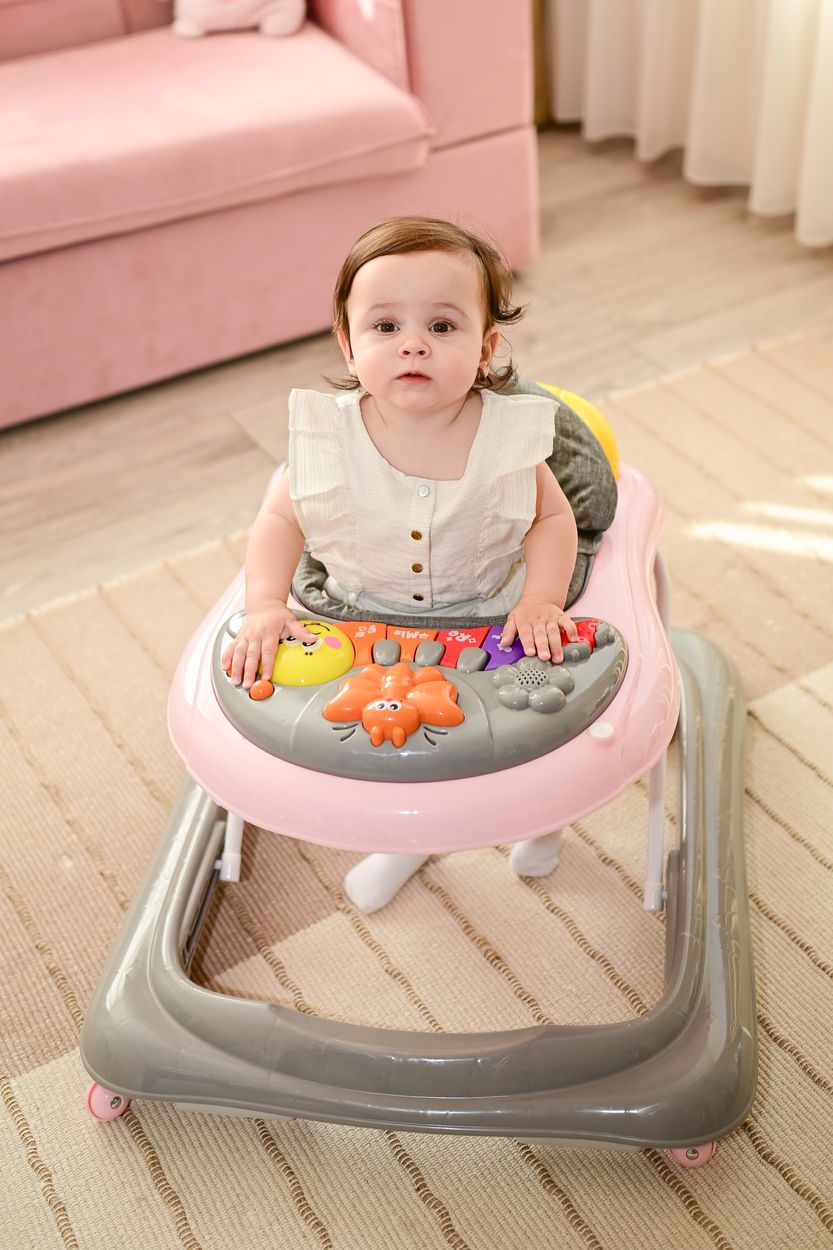 Ein Baby sitzt in einem rosa Lauflerner mit Spielzeug vor einem rosa Sofa.