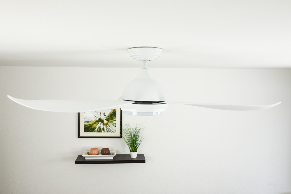 Ein weier Deckenventilator mit drei Blättern,-über einer kleinen schwarzen Regal mit Kunst und Pflanzen.