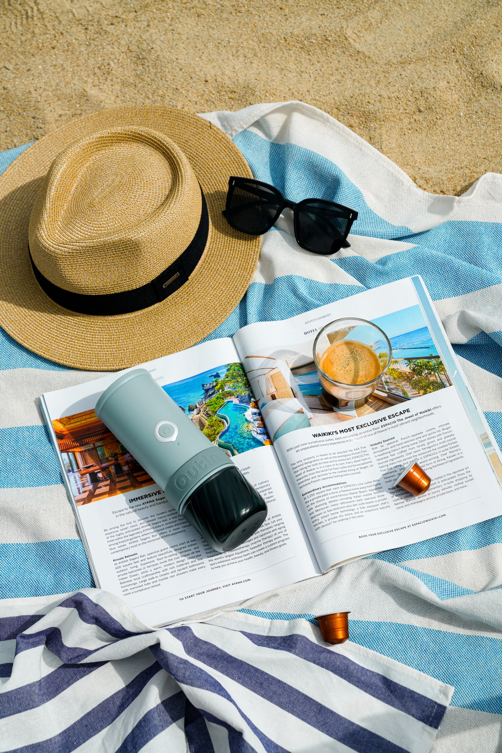 Un sombrero de paja, gafas de sol, una cafetera portátil y una revista sobre una toalla de playa.