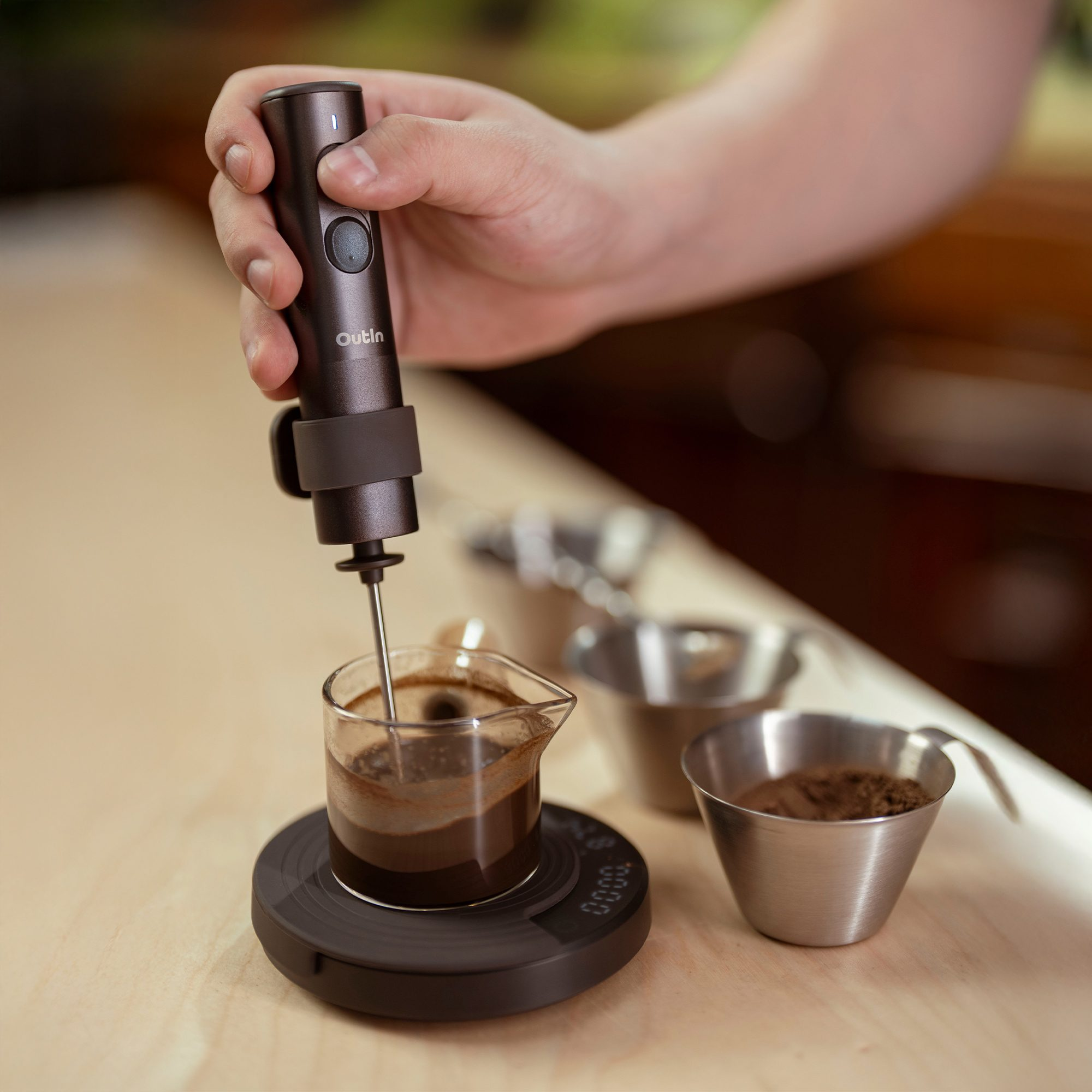 Una mano sostiene un espumador eléctrico marrón oscuro, mezclando café en un vaso de precipitados sobre una báscula.