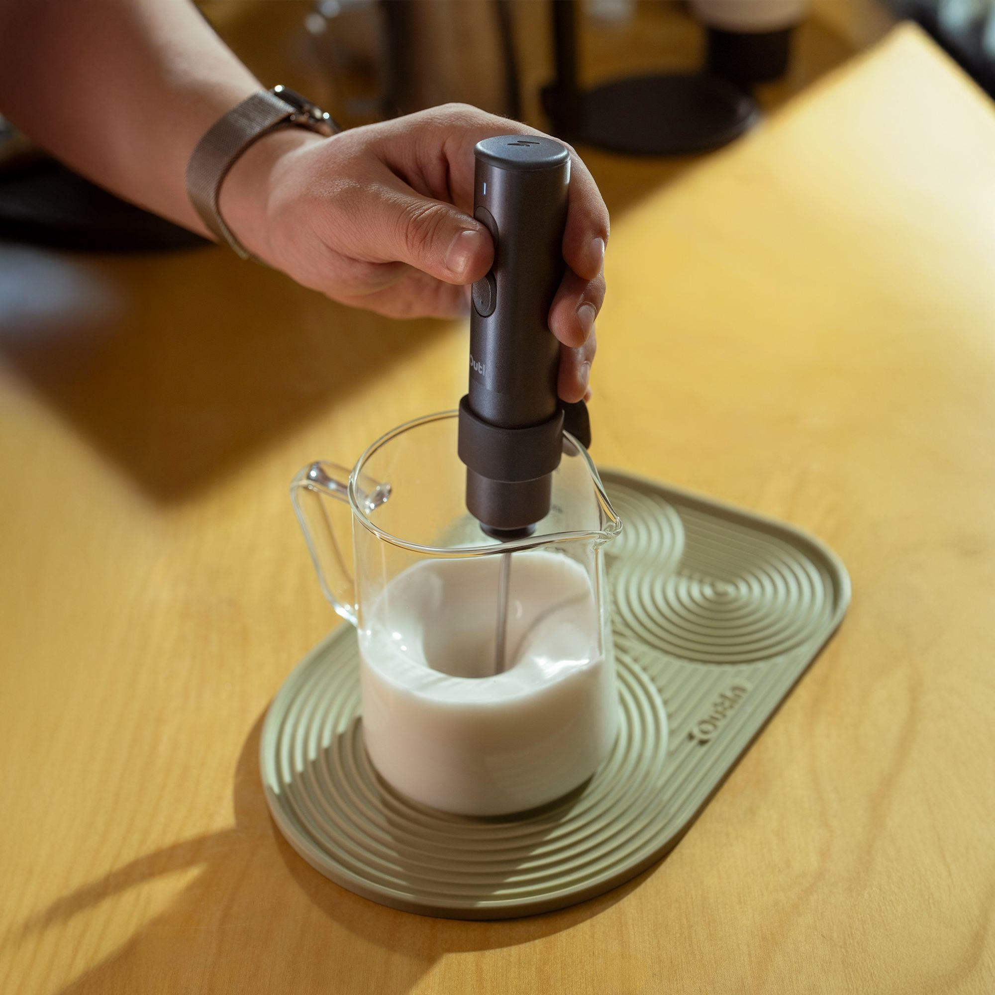 Una mano sostiene un espumador de leche oscuro sobre un vaso con líquido blanco.