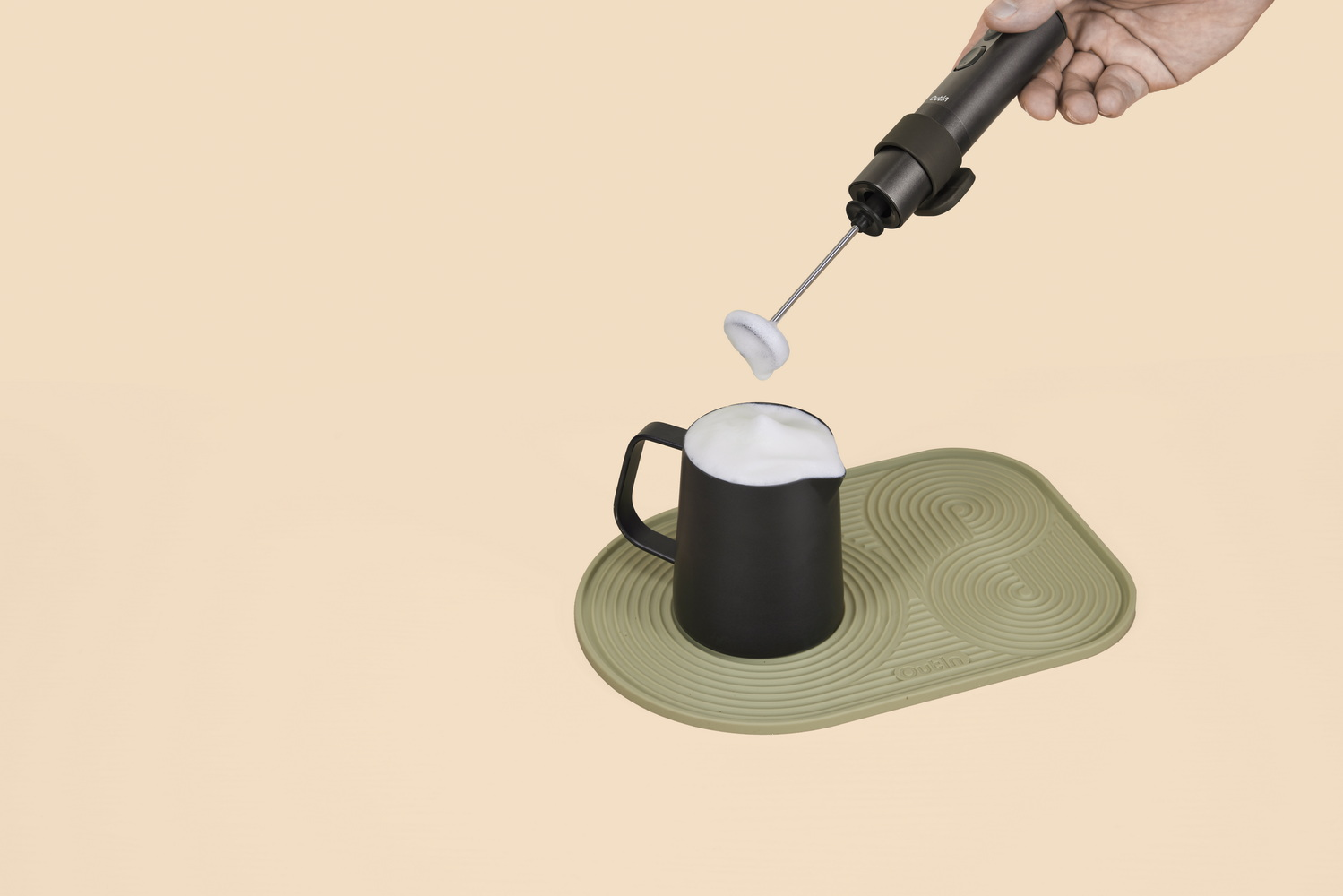 Mano sosteniendo espumador de leche sobre jarra negra con espuma sobre fondo beige.