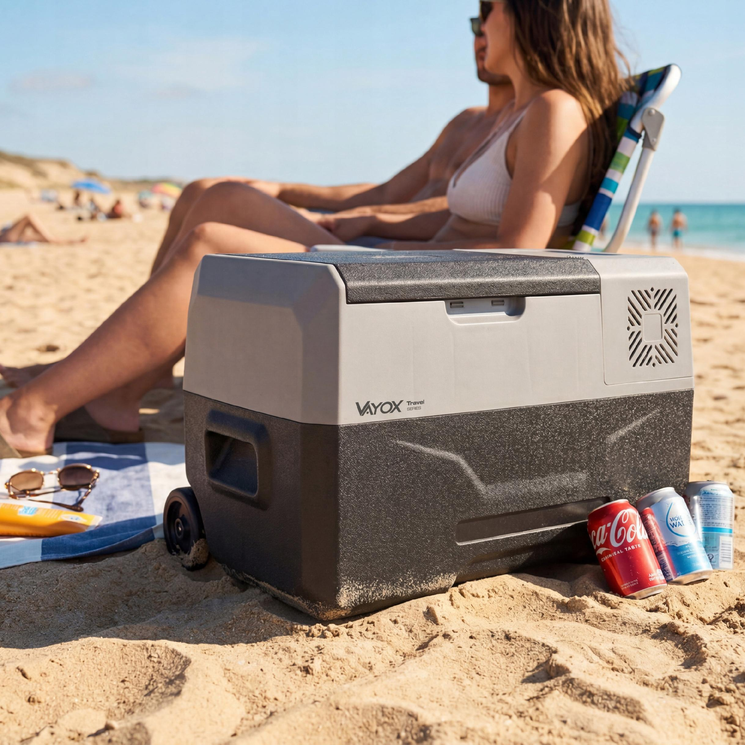 Ein tragbarer Kühler mit Getränkedosen am Strand, im Hintergrund ein Paar.