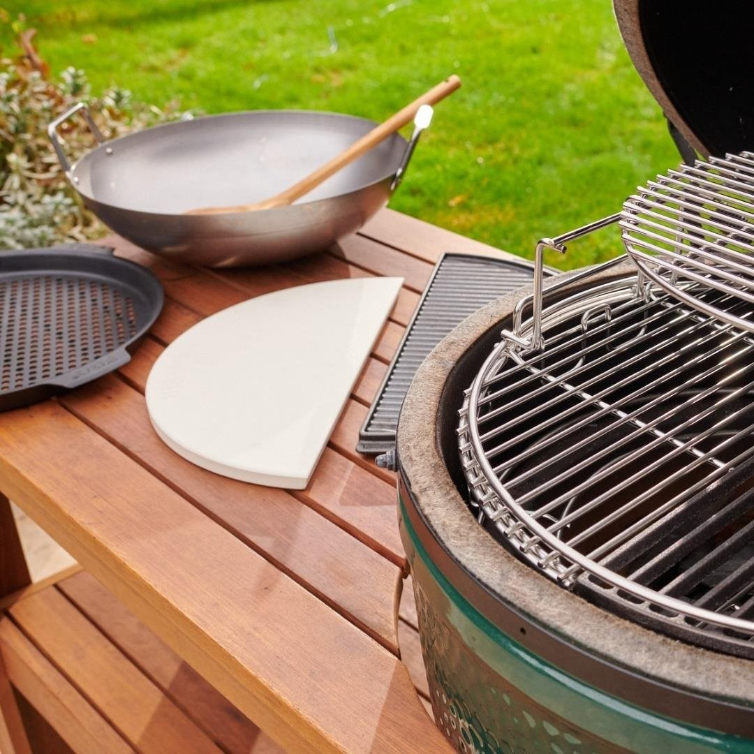 Ahşap masa üzerinde mangal aksesuarları: wok, pizza taşı, ızgara ve yeşil seramik mangal.