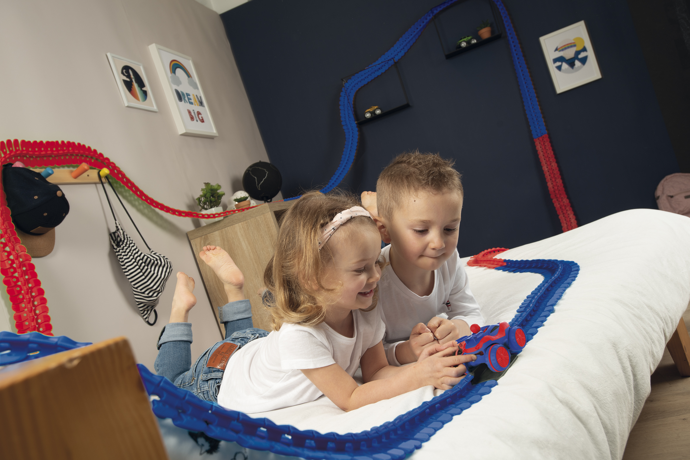 Due bambini giacciono su un letto, giocando con una pista per auto da corsa giocattolo blu.