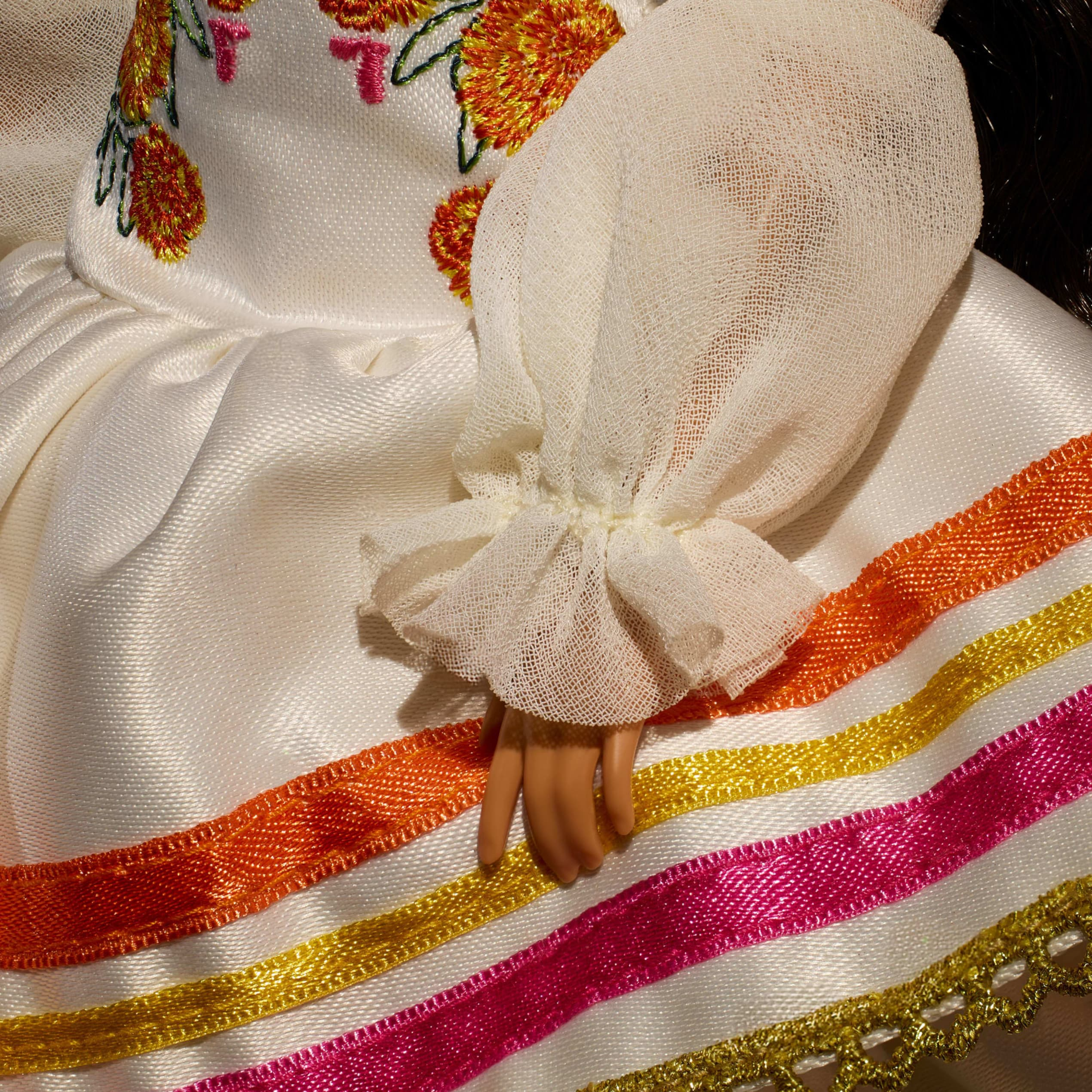 Detalle de muñeca con vestido blanco de satén, bordado floral y cintas coloridas.