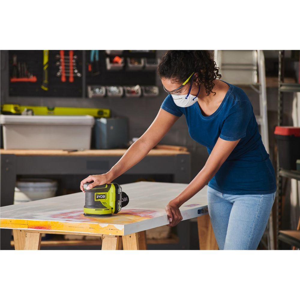 Frau schleift Tischplatte mit RYOBI-Schleifmaschine, trägt Schutzbrille und Maske.