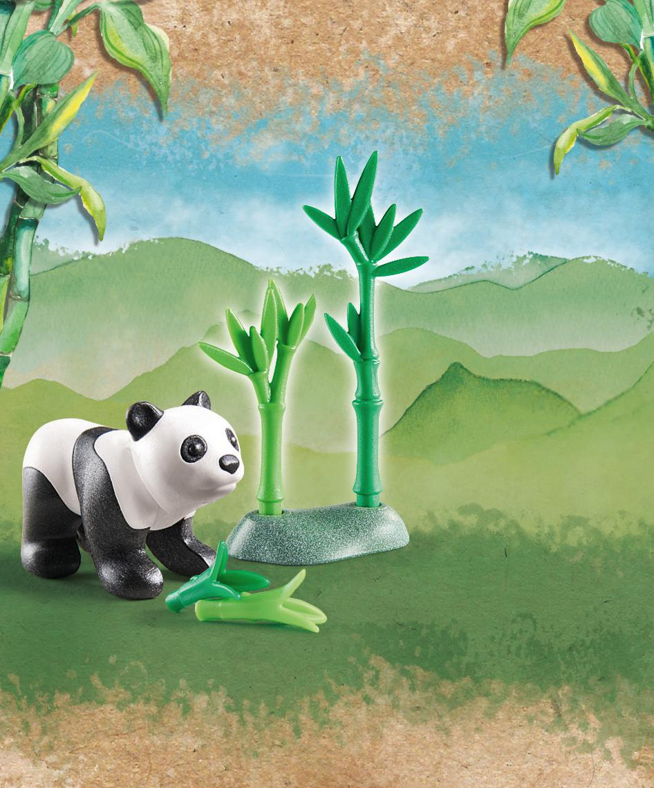 Ein Panda-Spielzeug steht auf grünem Boden mit zwei grünen Bambuspflanzen und herabgefallenen Bambusblättern, vor einer gemalten Berg- und Himmelskulisse.