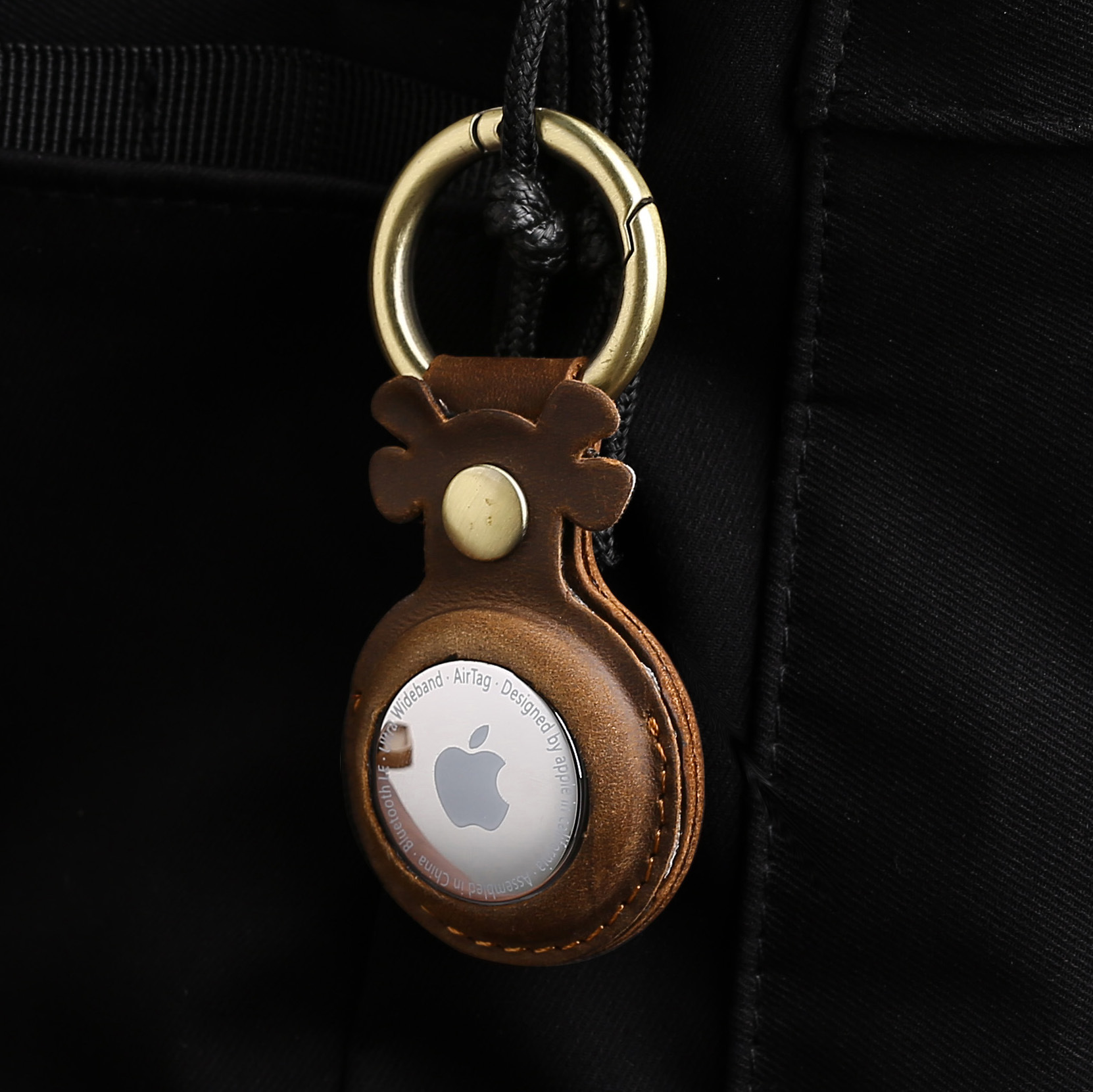 Brązowe skórzane etui na AirTag ze srebrnym logo Apple, przymocowane do czarnego plecaka.