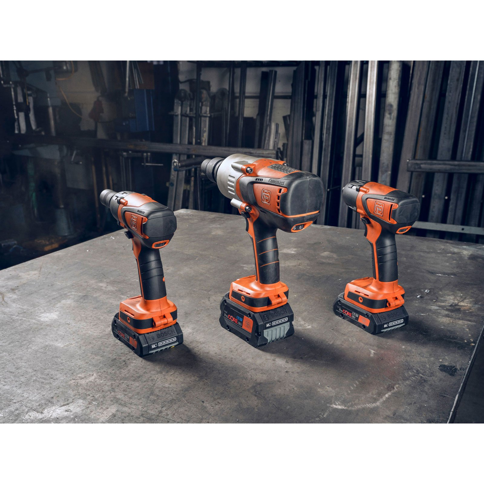 Drei orange-schwarze Akkuschrauber stehen auf einem Metalltisch. Im Hintergrund Metallstangen.