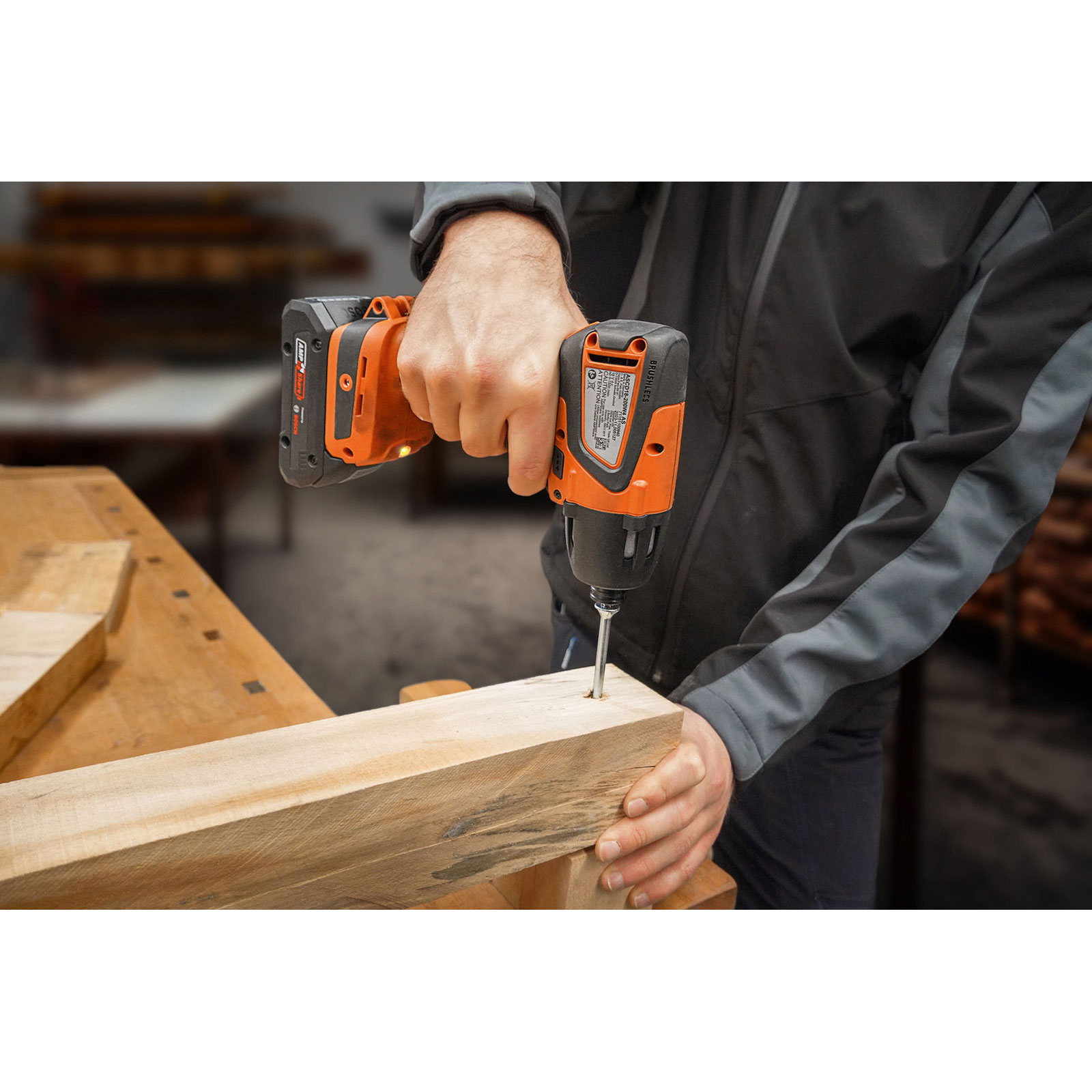 Eine Person benutzt einen orange-schwarzen Bohrer, um in eine Holzplanke zu schrauben. Werkstatt.