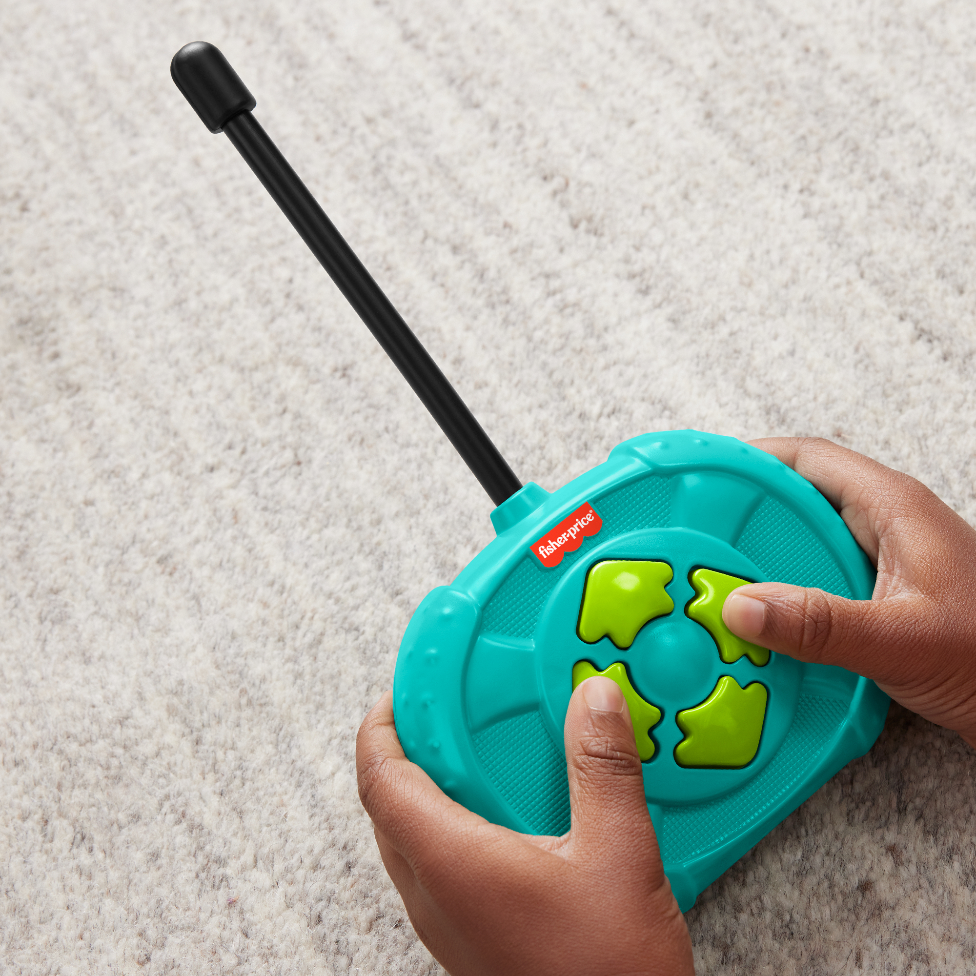 Manos sostienen un control remoto de juguete turquesa con botones verdes y antena negra sobre alfombra.