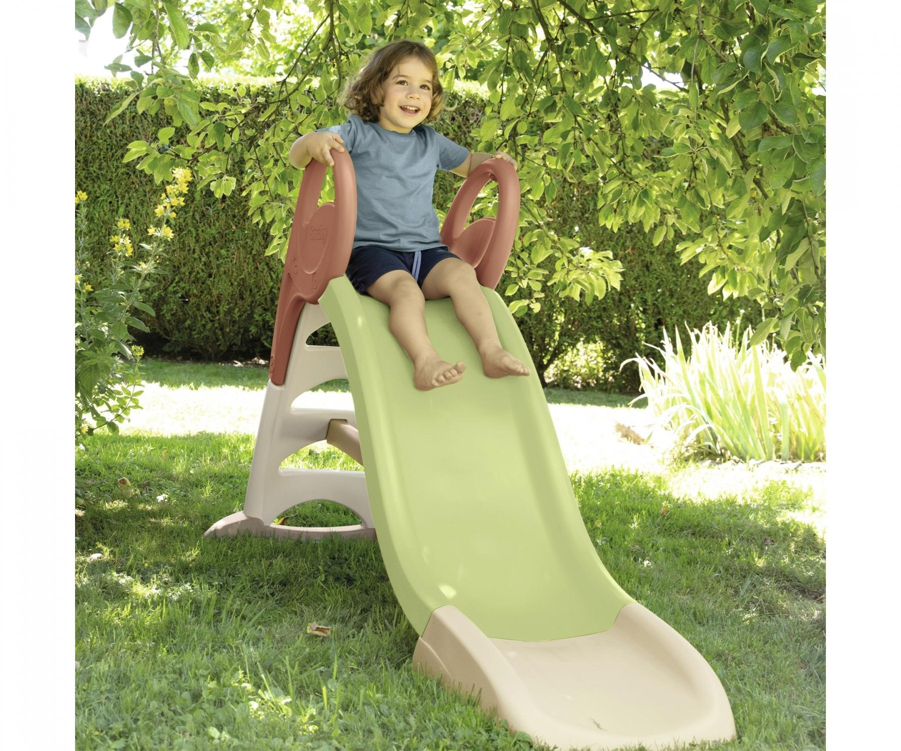 Un bambino siede su uno scivolo verde chiaro in un giardino, circondato da alberi.