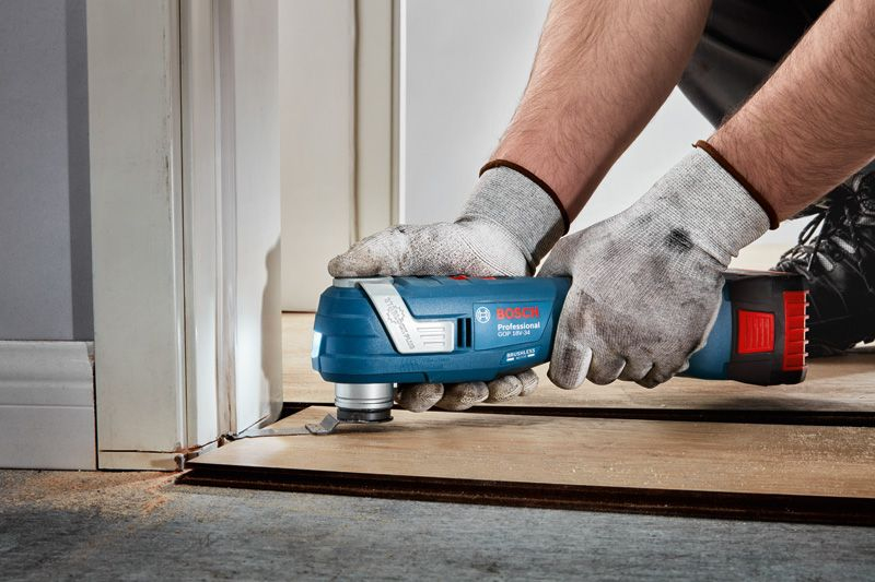 Mani guantate usano uno strumento oscillante Bosch blu per tagliare il parquet vicino a una porta.