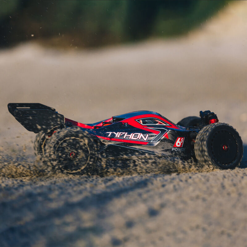 Un coche RC rojo y negro acelera sobre terreno arenoso, levantando polvo.
