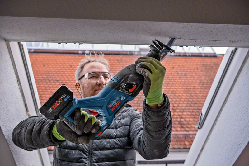 Mann mit Schutzbrille benutzt Säbelsäge an Dachfenster, rote Ziegeldächer im Hintergrund.