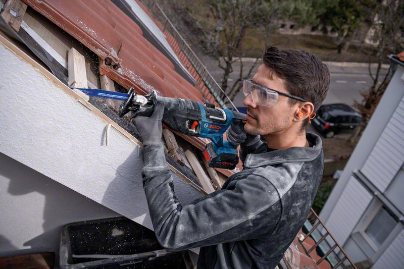 Mann mit Schutzbrille und Gehörschutz sägt auf einem Dach mit Säbelsäge.