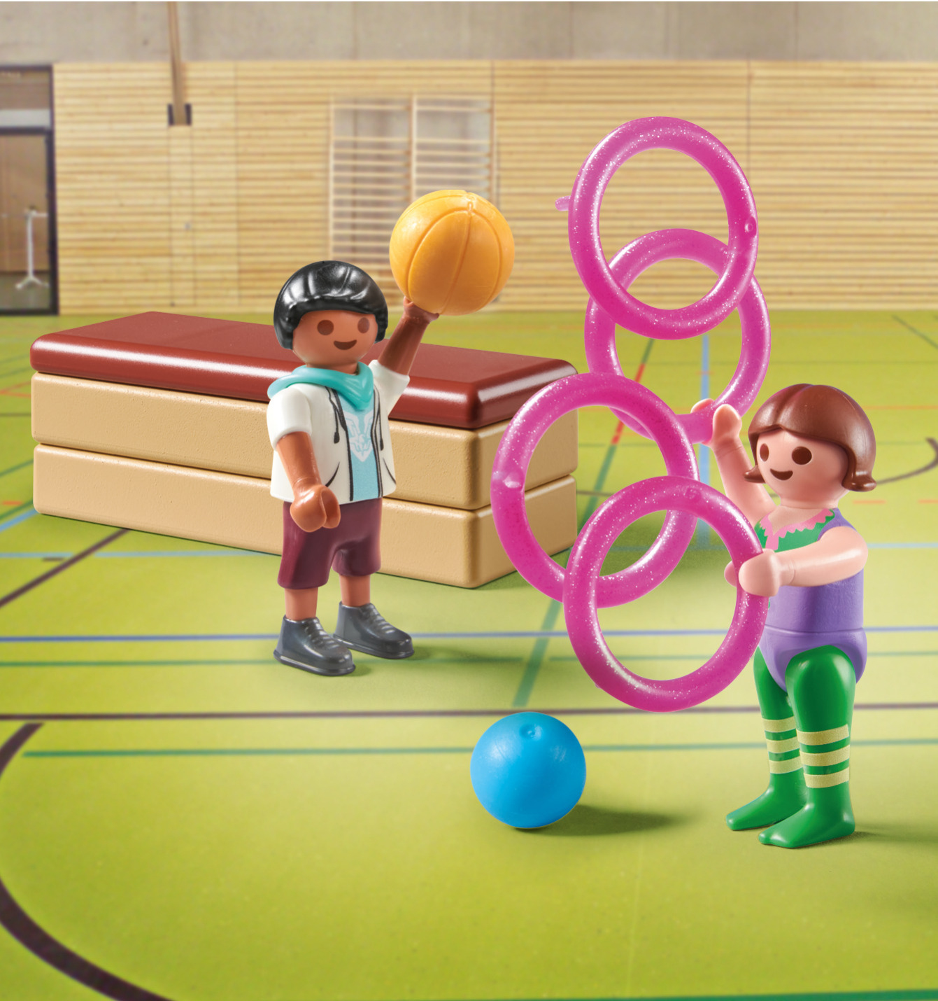 Zwei Kinder in einer Turnhalle mit Sportgeräten: Basketball, Ringe und ein blauer Ball.