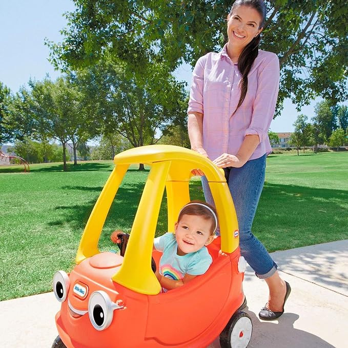 Una donna spinge un bambino in un'auto giocattolo rossa e gialla su un sentiero lastricato in un parco con erba verde e alberi.