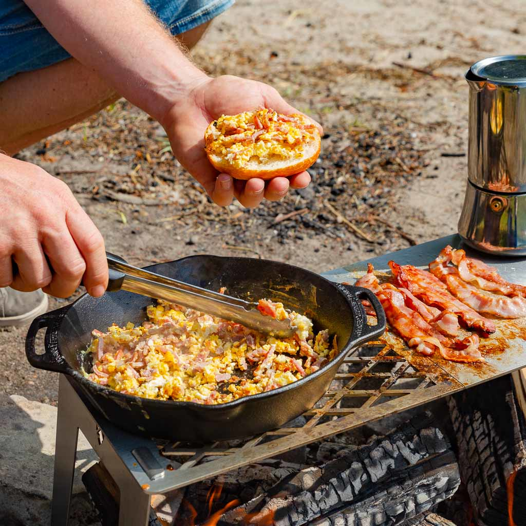 Ein Mann bereitet Rührei mit Speck über einem Lagerfeuer zu und füllt ein Brötchen.