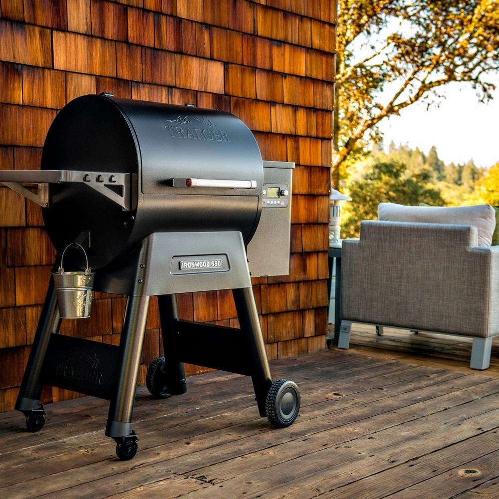 Una parrilla Traeger negra se asienta en una terraza de madera contra una pared revestida de madera.