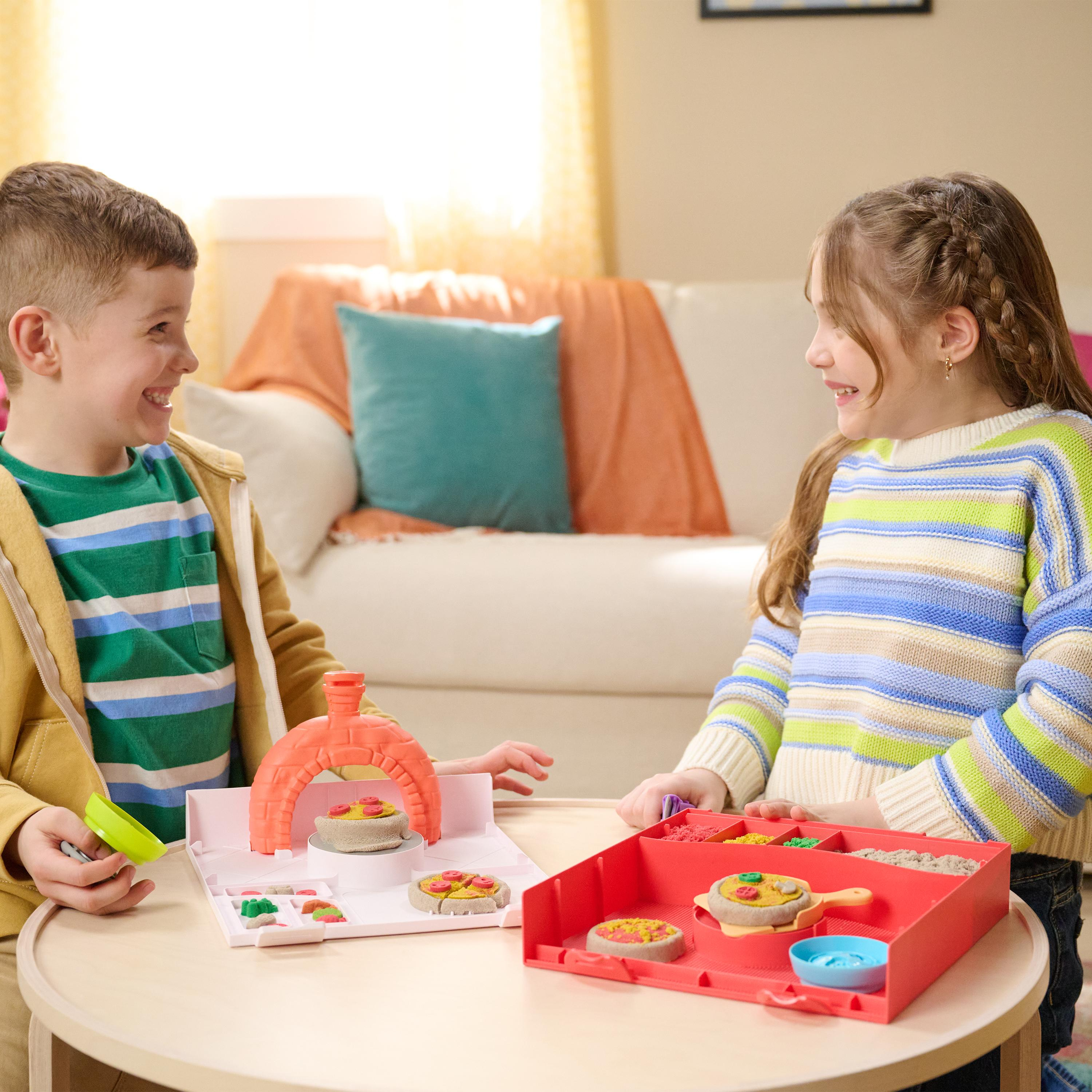 Dos niños, un niño y una niña, se sonríen mientras juegan con un juego de hacer pizzas en una mesa de madera.