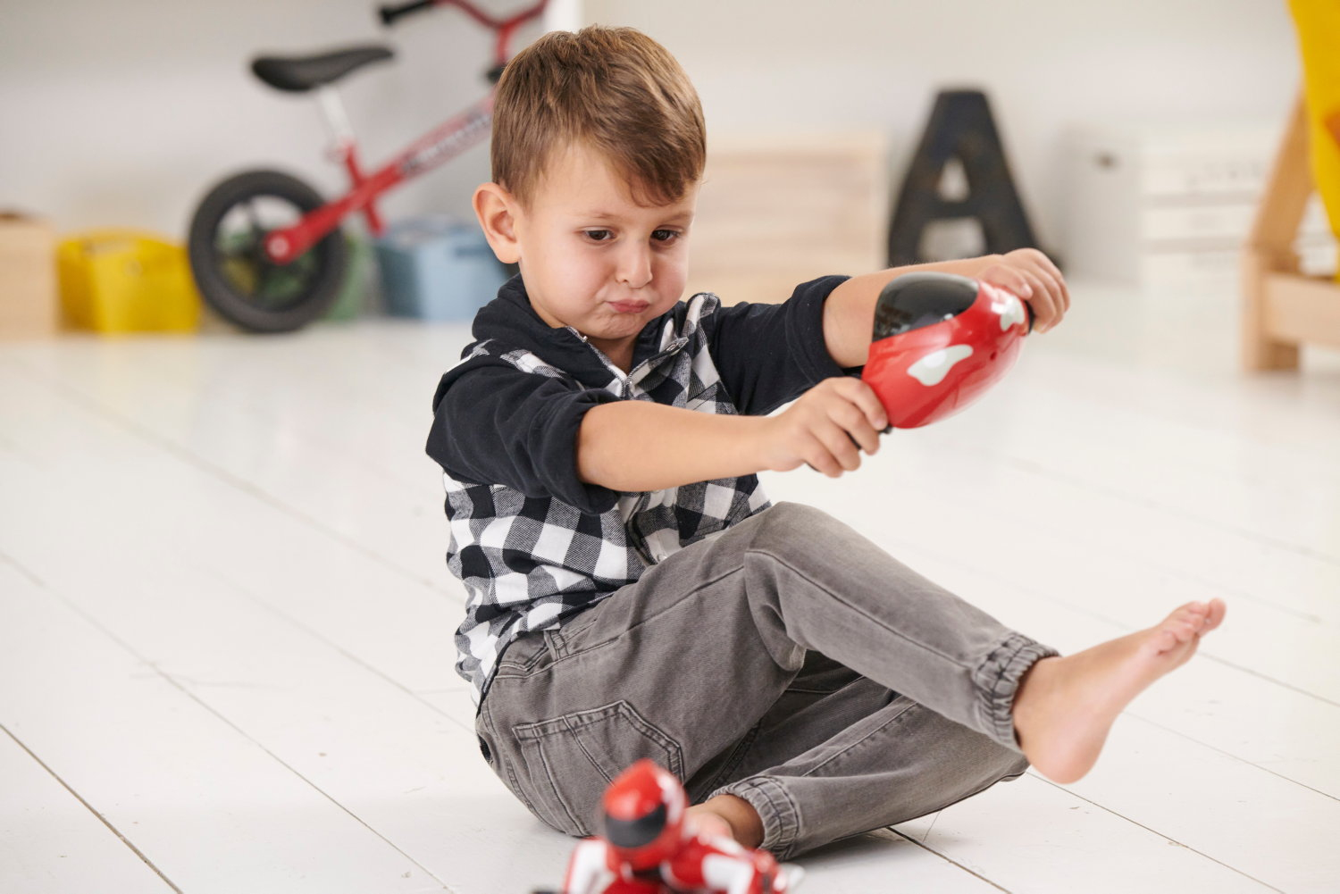 Un niño sentado en el suelo jugando con un coche de juguete rojo. Una bicicleta de equilibrio al fondo.