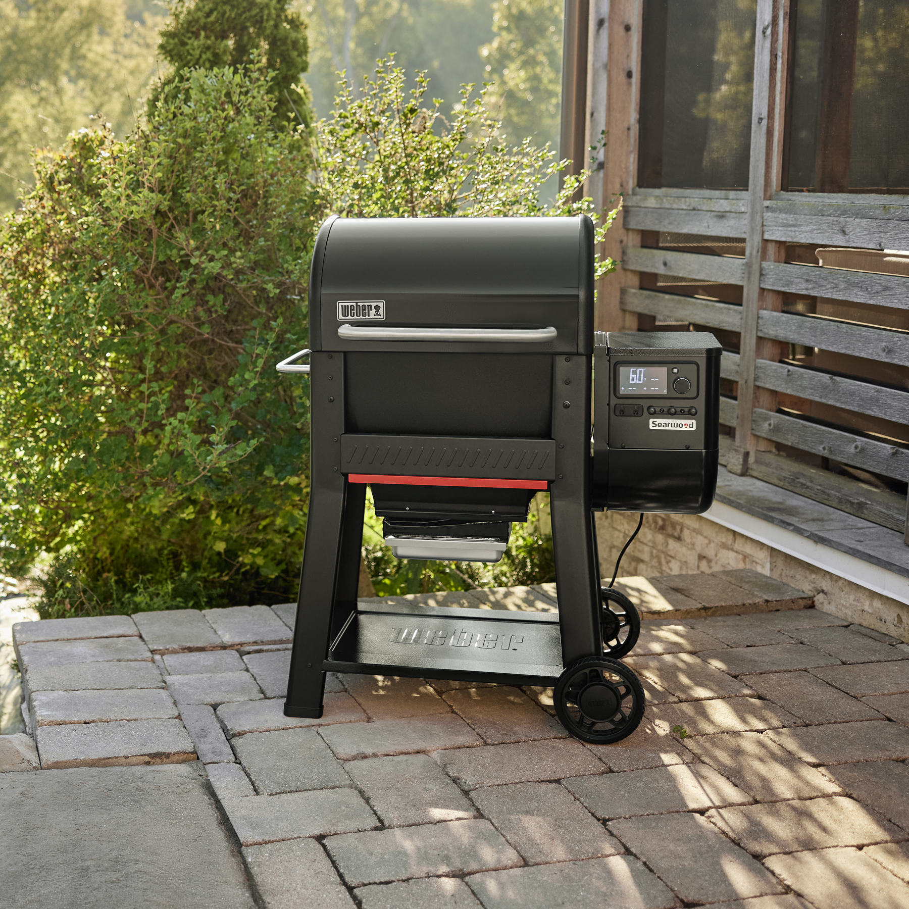 Una griglia Weber nera si trova su un patio pavimentato, con struttura in legno e vegetazione sullo sfondo.