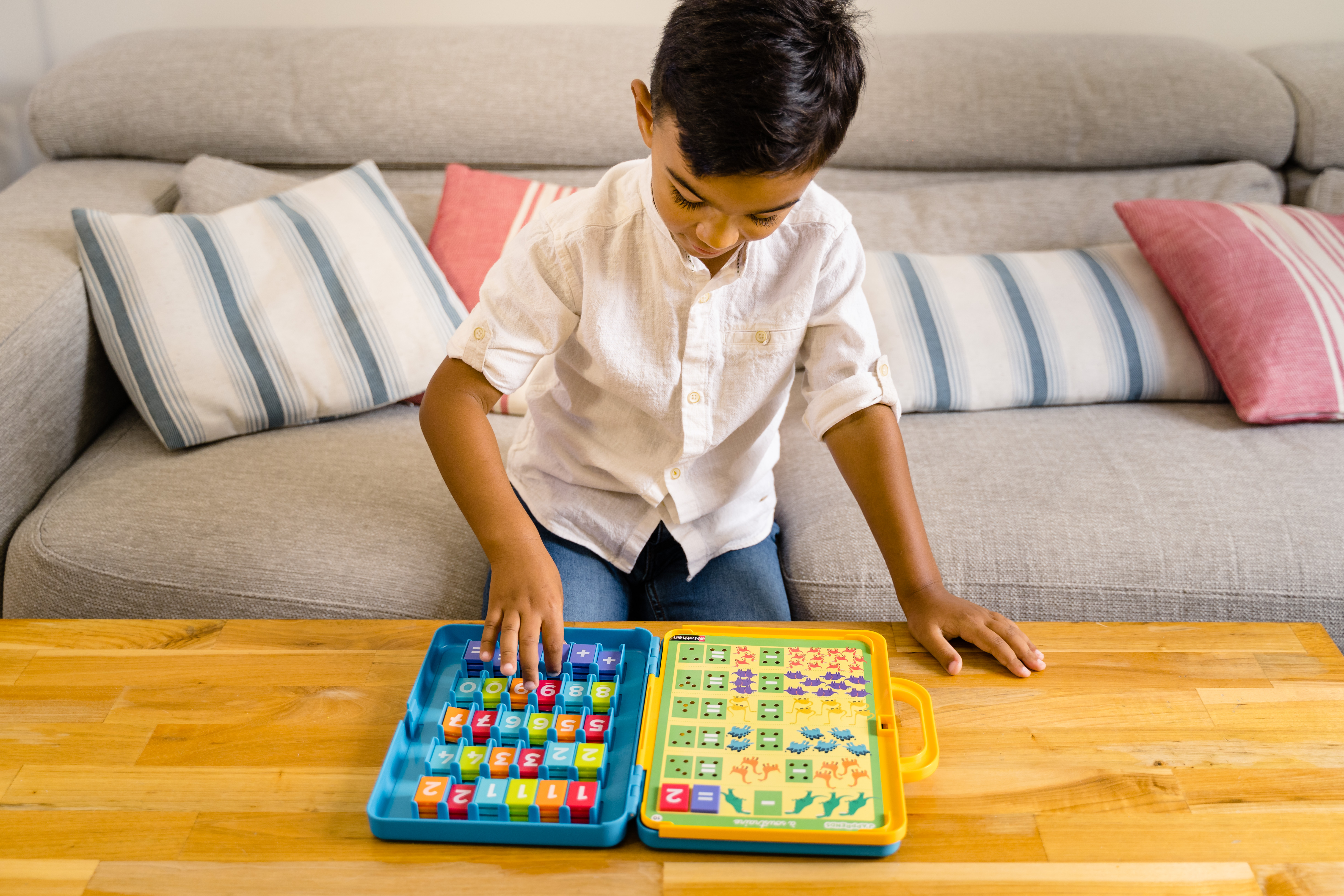 Ein kleiner Junge, gekleidet in ein weißes Hemd und blaue Jeans, sitzt auf einem grauen Sofa und spielt mit einem Zahlenspielzeug.
