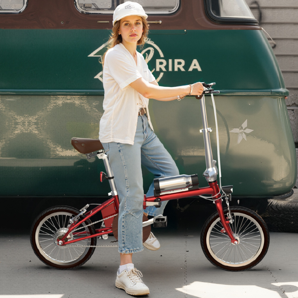 Eine Frau mit weißer Kappe und weißem Hemd sitzt auf einem roten Klapprad vor einem grünen Fahrzeug.