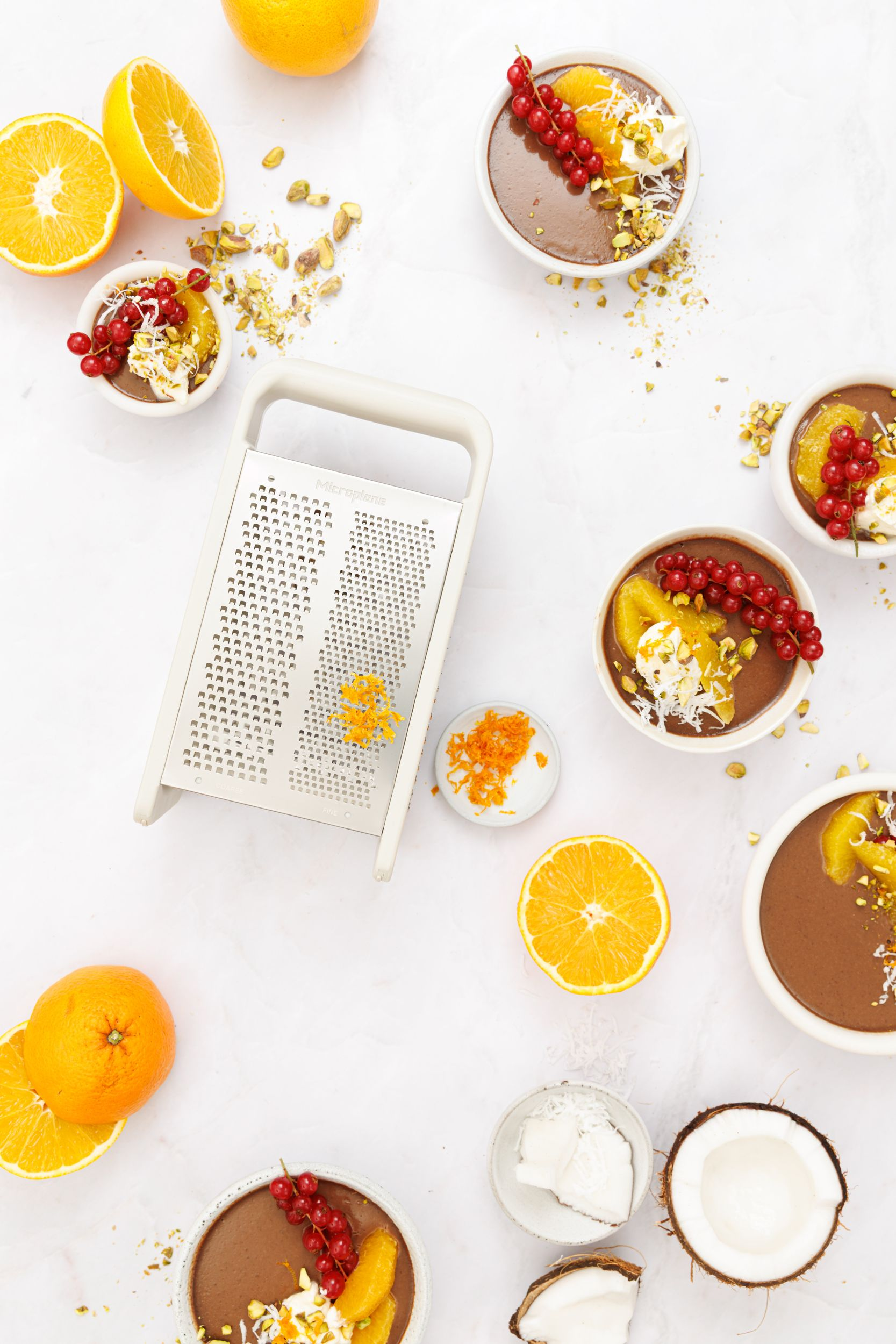 Draufsicht auf Schokoladenpudding, Orangen, Kokosnüsse und eine Reibe auf einer weißen Oberfläche.