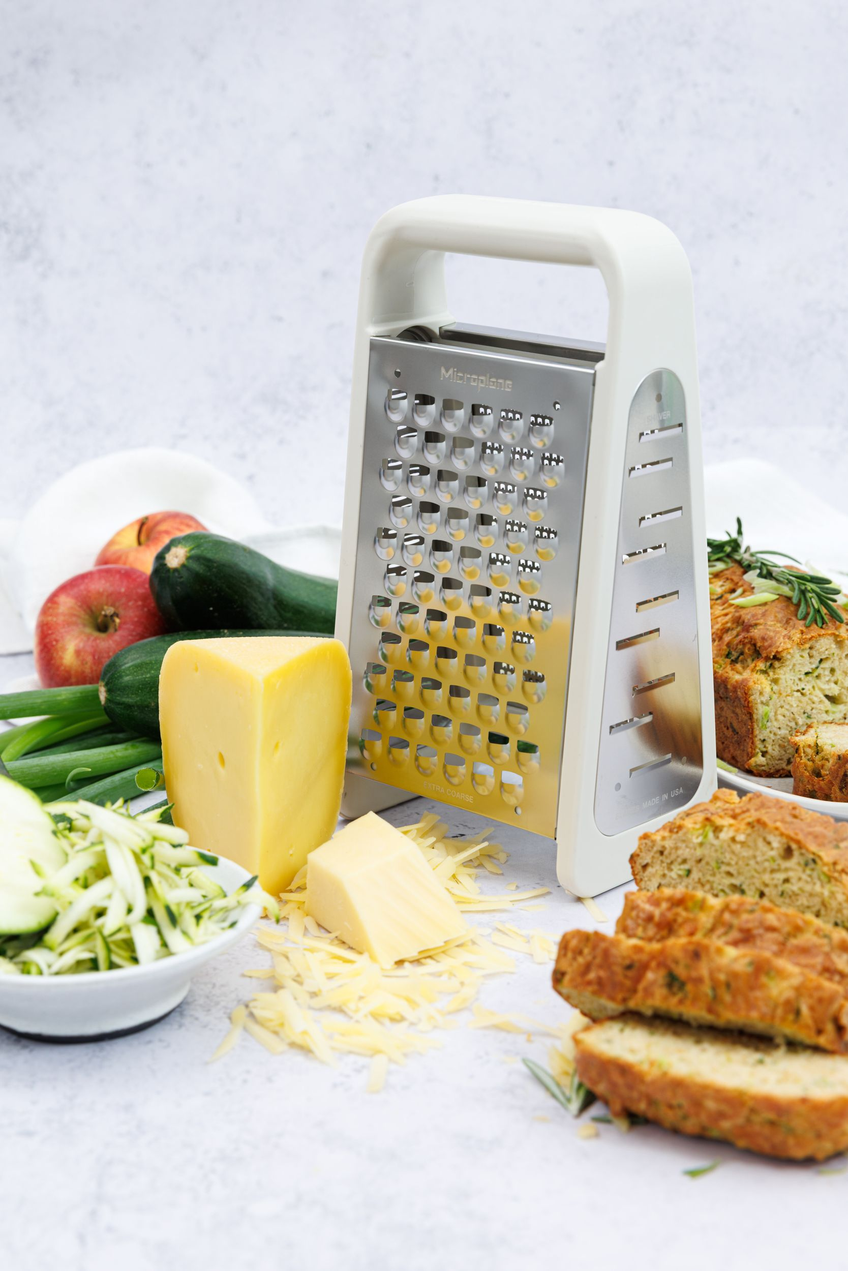 Ein Handreibeisen steht neben Zucchini, Äpfeln, Käse und Brot.