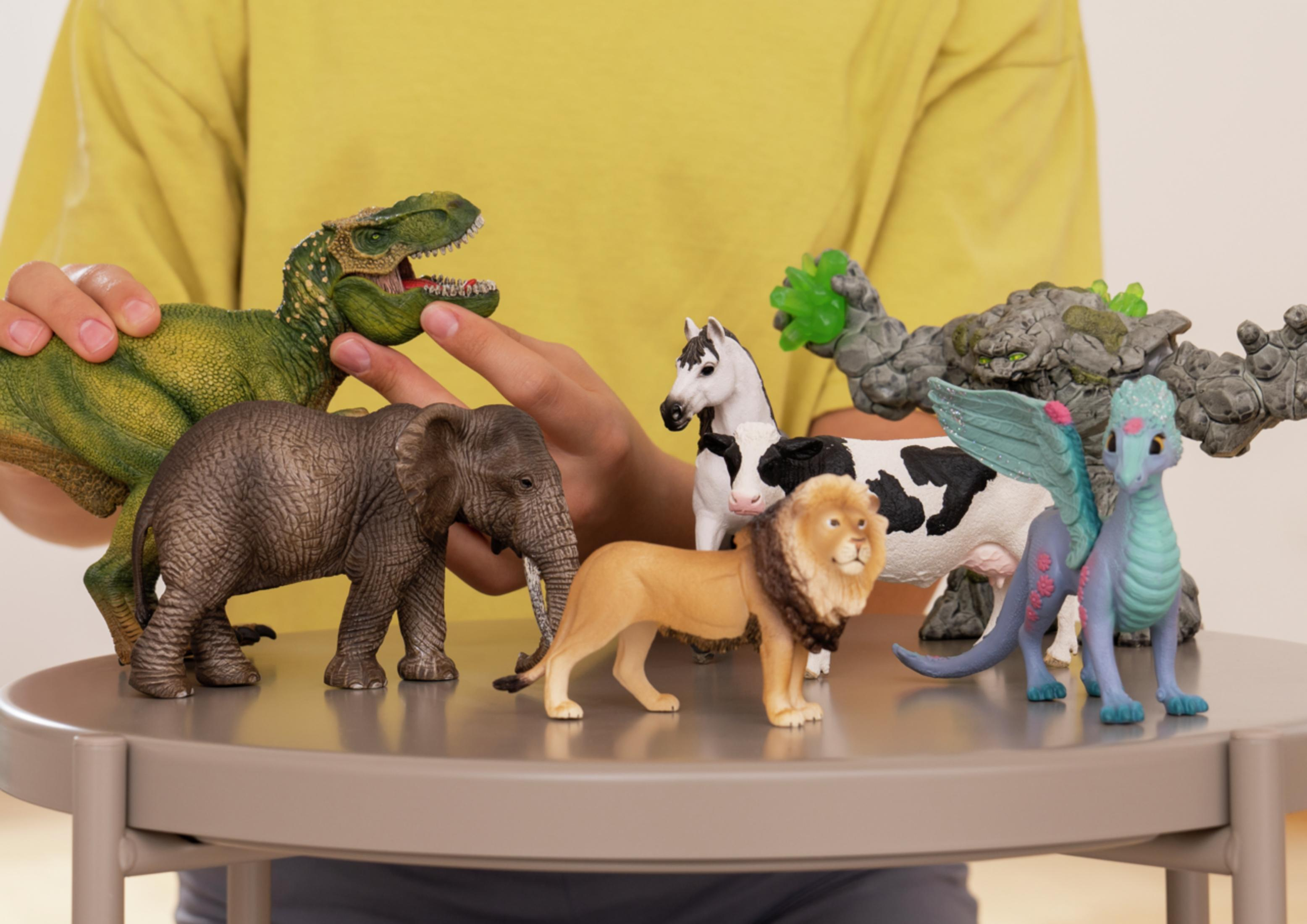 Een hand houdt een groen dinosaurus speelgoed vast boven een olifant.