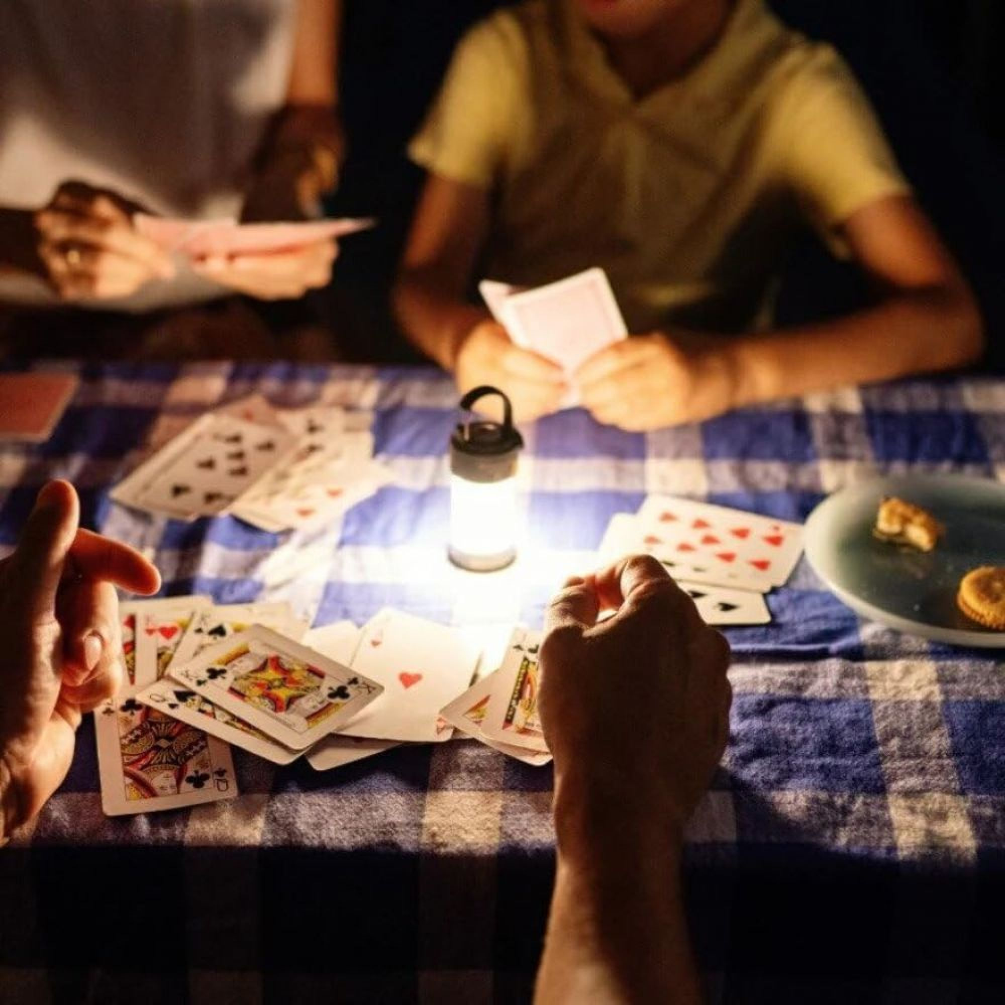 Drei Personen spielen Karten bei Laternenlicht auf einem blau-weiß karierten Tischtuch. Hände halten Karten, weitere sind verstreut.