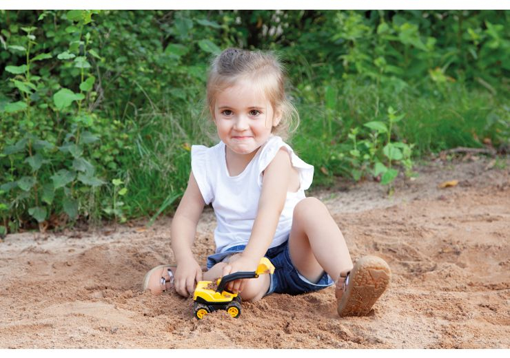 Ein kleines Mädchen sitzt im Sand und spielt mit einem gelben Spielzeugbagger.