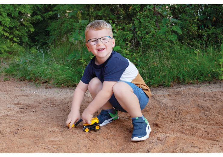 Ein Junge mit Brille spielt mit einem gelben Spielzeugbagger im Sand.