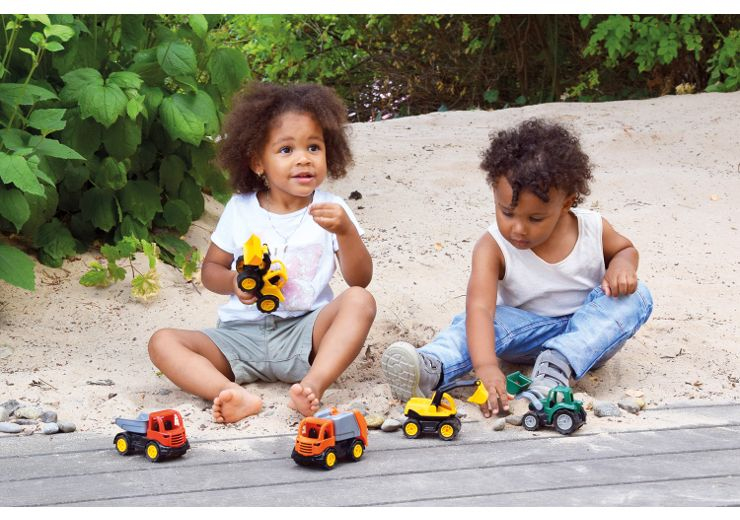 Zwei Kleinkinder spielen mit Spielzeug-Baufahrzeugen auf sandigem und holzigem Untergrund, umgeben von grünem Laub.