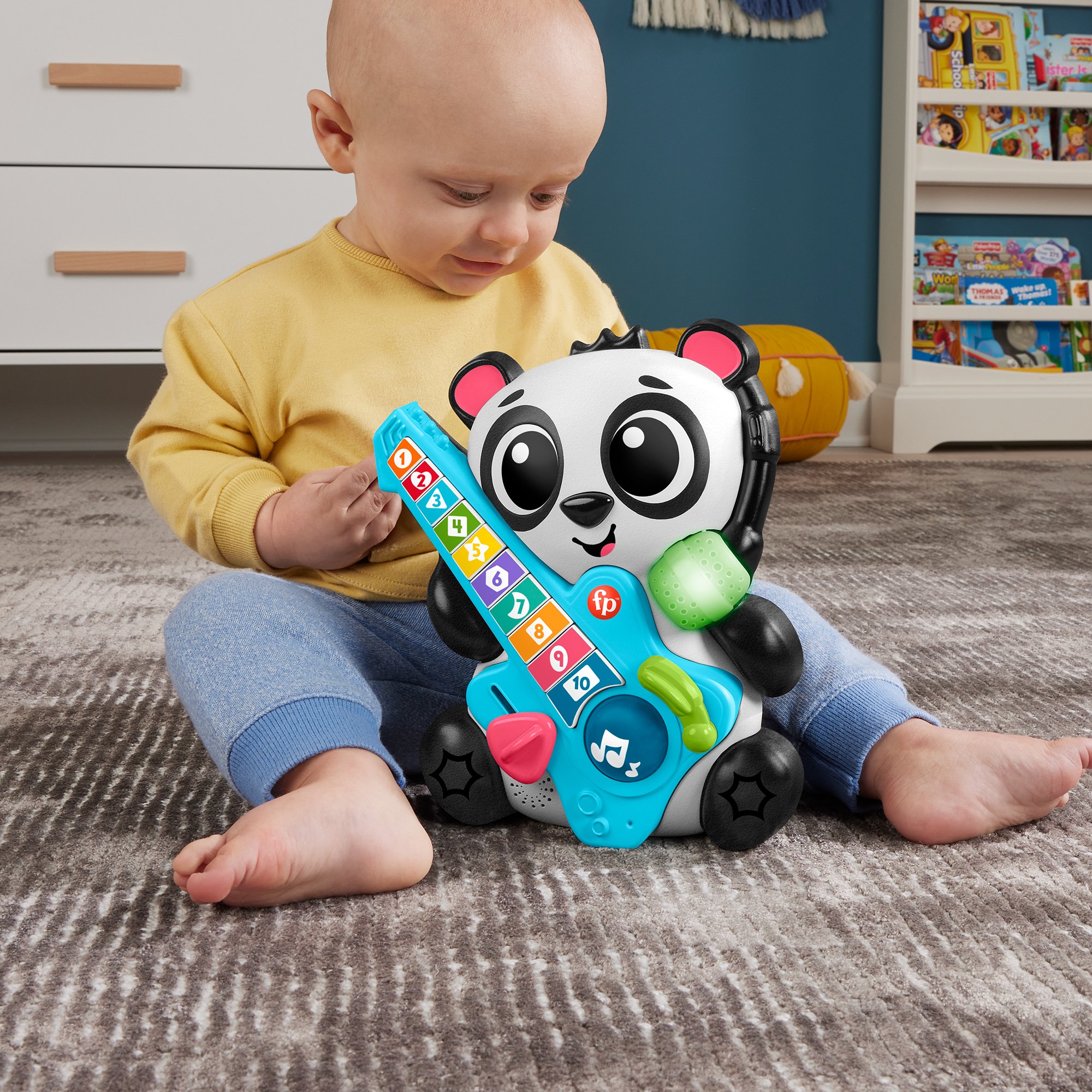 Ein Baby in gelbem Oberteil und blauer Hose sitzt auf einem Teppich und spielt mit einem schwarz-weißen Panda-Gitarrenspielzeug.