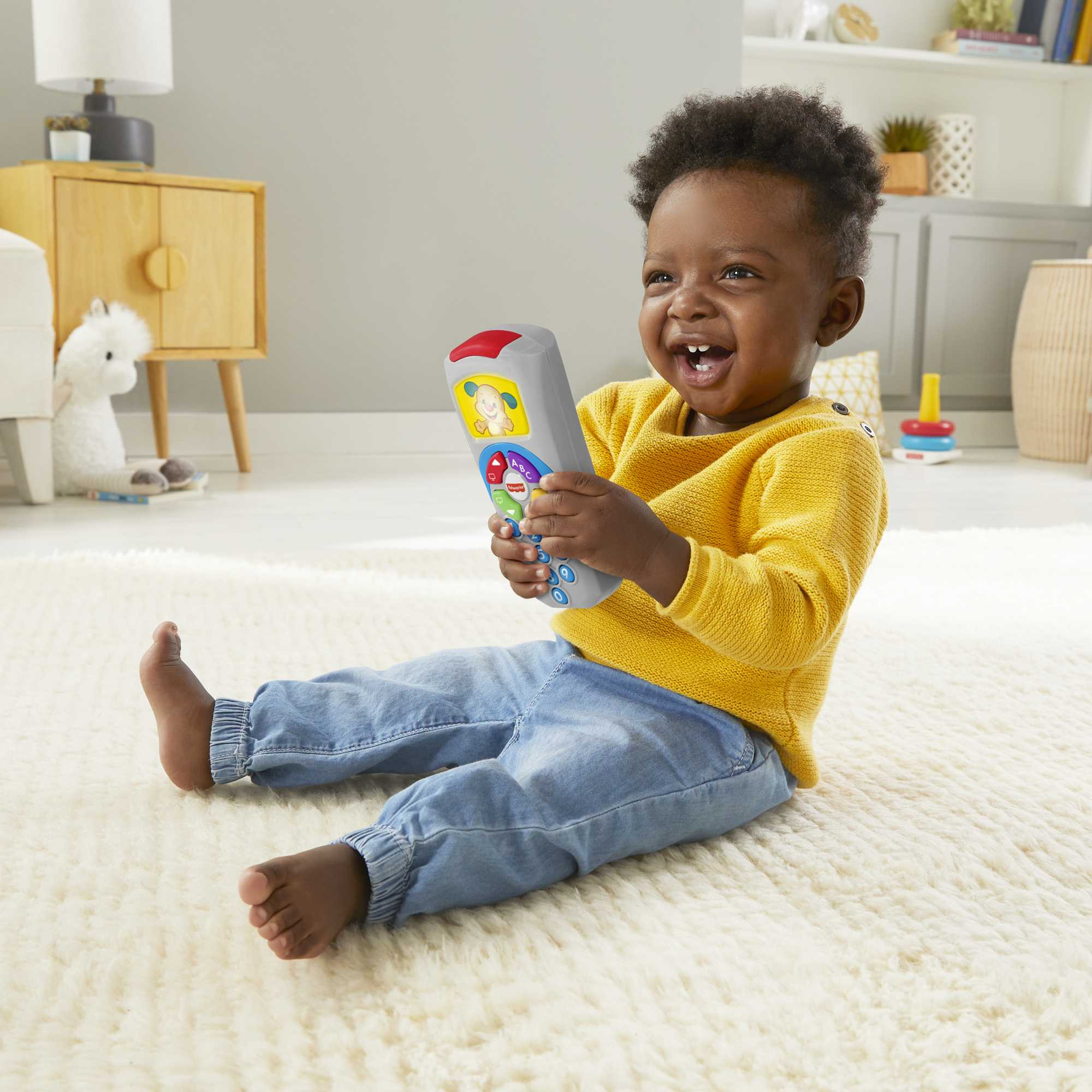 Ein Baby in einem gelben Pullover spielt mit einer Fernbedienung auf einem cremefarbenen Teppich.