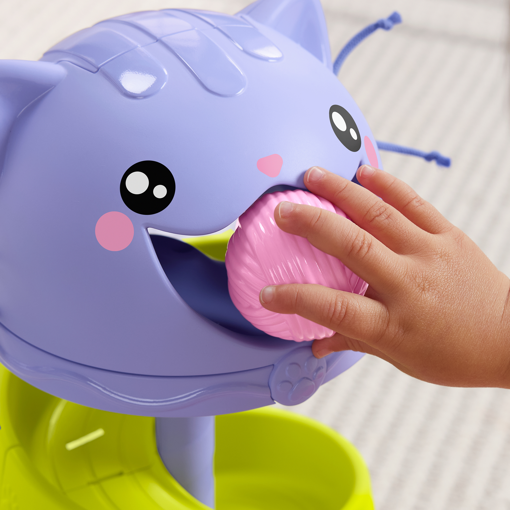 Ein Kinderfinger steckt einen rosa Ball in das Maul eines lila Katzenkopfspielzeugs.