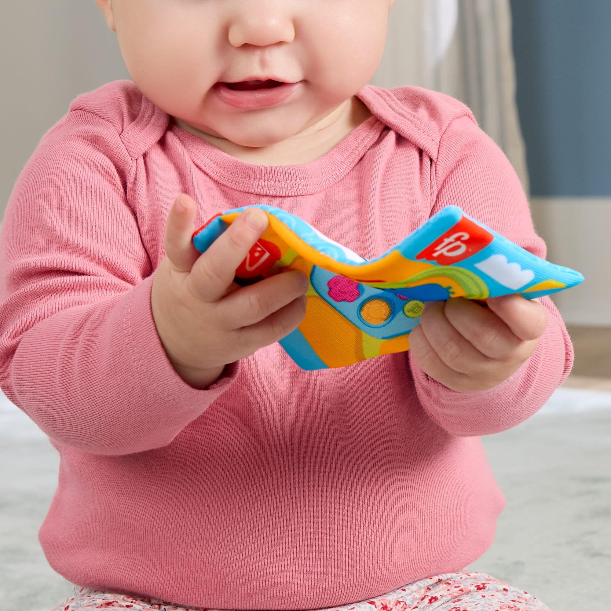 Ein Baby in einem rosafarbenen Strampler hält ein buntes Stoffspielzeug.
