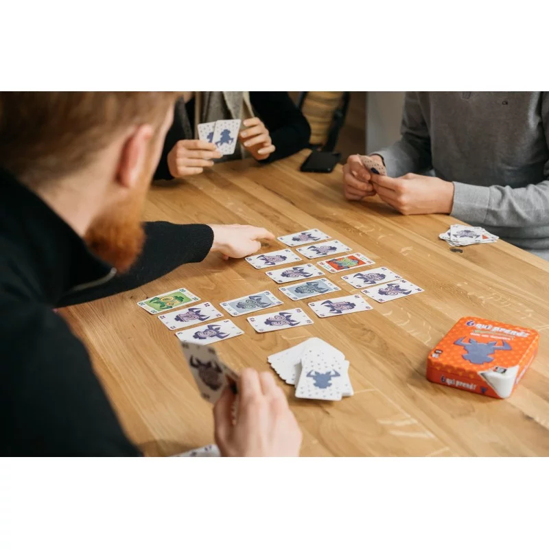 Trois personnes jouent à un jeu de cartes sur une table en bois, avec des cartes étalées et des mains tenant des cartes.